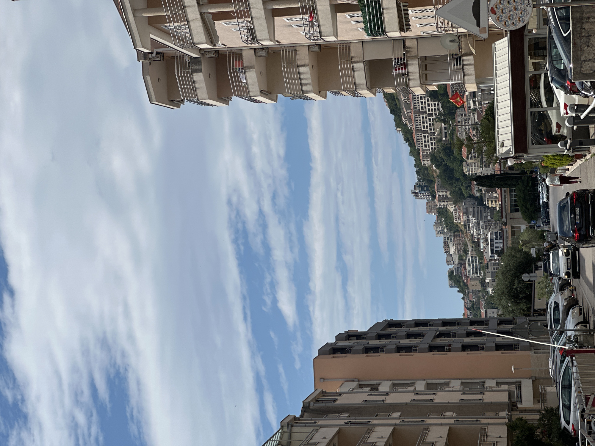 The image depicts a street scene in Budva, Montenegro, with buildings lining both sides of the road. The sky above is partly cloudy. The buildings on the left feature multiple balconies. On the right side of the street, a row of parked cars and a few people are visible. In the background, a densely built hillside with various buildings can be seen.