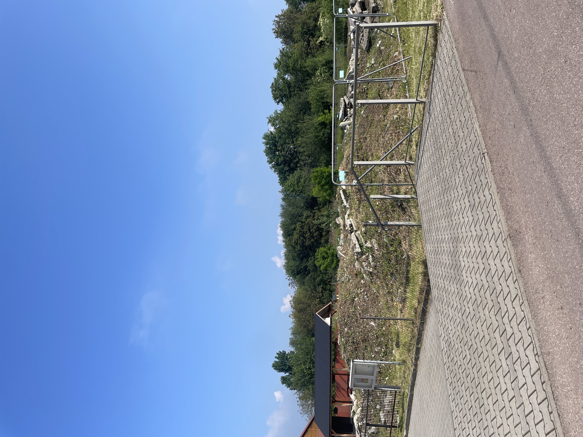 An outdoor scene is depicted under a bright, clear blue sky with only a few faint white clouds. A dense line of green trees and foliage spans the horizon, suggesting a wooded area.
In the middle ground, directly in front of the trees, lies a grassy and dirt area strewn with scattered rocks or debris. This section is cordoned off by a long metal barrier, consisting of silver-colored posts and horizontal rails, with some green elements attached. The barrier runs diagonally across the frame.
In the foreground, a paved path made of grey interlocking bricks is visible, running parallel to the metal barrier. Adjacent to this paved path on the right is a dark asphalt or road surface. On the far left of the image, part of a wooden building with a dark roof and reddish-brown walls is visible, nestled among trees. A white information board with an unreadable text and a black frame stands near this building, accompanied by a small black metal fence.
The image captures a static, calm, and well-lit daytime scene, indicating fair weather. No individuals or specific activities are observable. The setting appears to be a public outdoor space, possibly a park or an area near an archaeological site or undergoing development, given the debris and barriers.