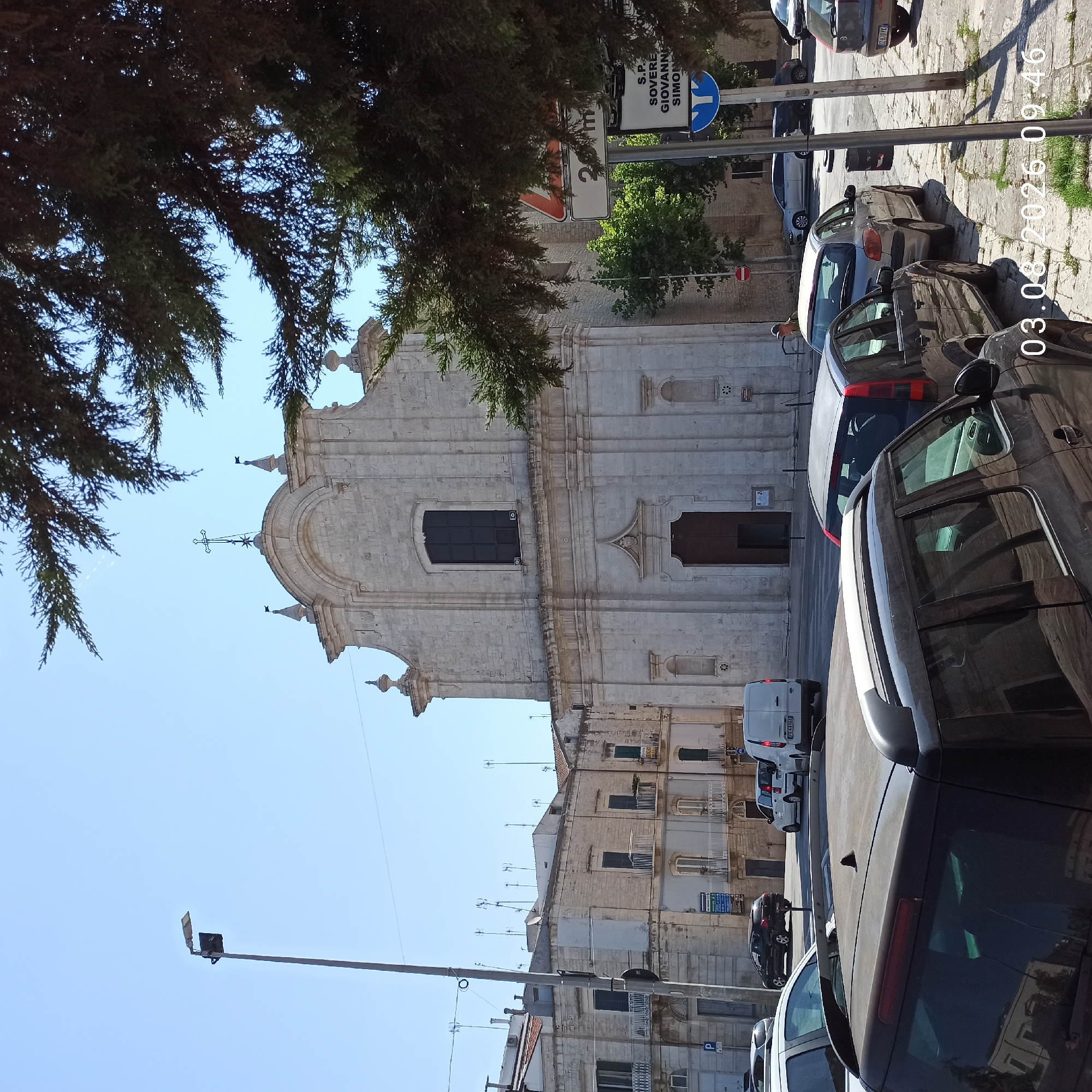 The image captures an outdoor scene in Ruvo di Puglia, Italy, featuring a church and surrounding urban environment. In the foreground, several parked cars are visible, with a dark-colored SUV dominating the lower left portion of the frame. The mid-ground shows a white van, a silver hatchback, and a black SUV. The background is dominated by a large, light-colored stone church with a prominent arched entrance and a dark window above. The church facade exhibits classical architectural details. To the left of the church, several multi-story buildings with a classical architectural style are visible, displaying balconies and windows. A street lamp with a light fixture is positioned on the left side of the image. Signs are present on the right side of the frame, including a traffic sign indicating "20 m" and another sign with Italian text, "S.P. SOVERE GIOVANNI SIMON". The sky is clear and blue. The ground is paved with cobblestones in some areas and concrete in others, with patches of grass. The overall lighting suggests a bright, sunny day. A date and time stamp "03.06.2026 09:46" is superimposed on the lower right corner of the image.