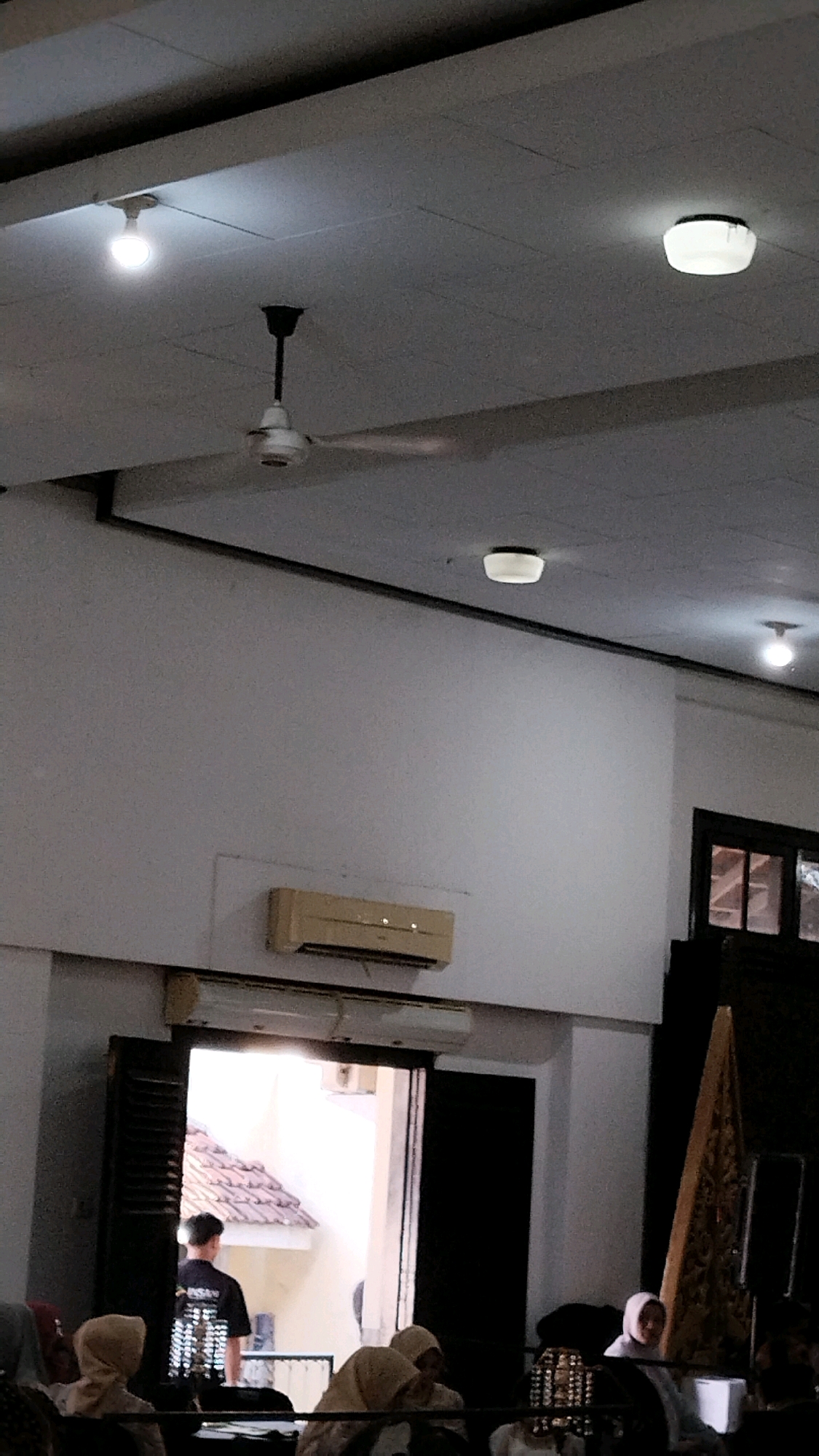 The image shows the interior of a room, possibly a hall or meeting space, with a high ceiling. Several light fixtures are visible on the ceiling, including a white bulb on the left, a round flush-mount light in the upper right, and another light fixture further to the right. A white ceiling fan with black blades is also mounted on the ceiling.
Below the ceiling, a light-colored air conditioning unit is mounted on the wall. In the lower part of the image, a doorway is open, revealing a view of an outdoor area with a tiled roof. A person wearing a black t-shirt with white print is standing in the doorway, facing away from the camera.
In the foreground, several people are seated, mostly wearing headscarves. On the right side of the image, a tall, triangular decorative item, possibly a traditional puppet or screen, is visible. The overall lighting suggests it is either evening or the interior lights are on, creating a somewhat dim atmosphere. The scene appears to be a gathering or event. The presence of a ceiling fan and air conditioning unit suggests a warm climate, consistent with Jember, Indonesia.