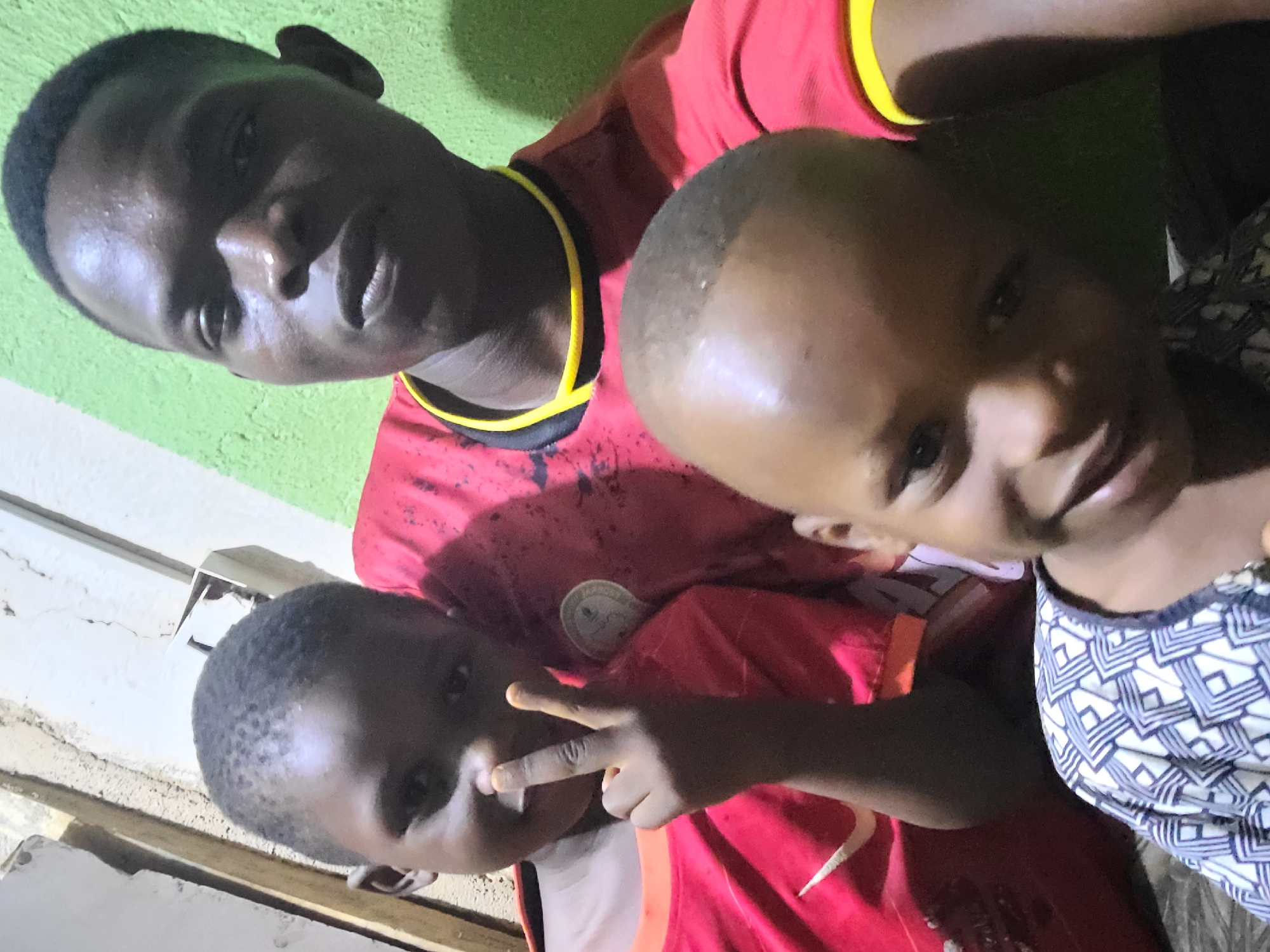 The image shows three individuals, possibly family members, posing close together in what appears to be an indoor setting. The background features a wall that transitions from green to white. One of the children is holding up a peace sign. The location is indicated as Unknown, Unknown.