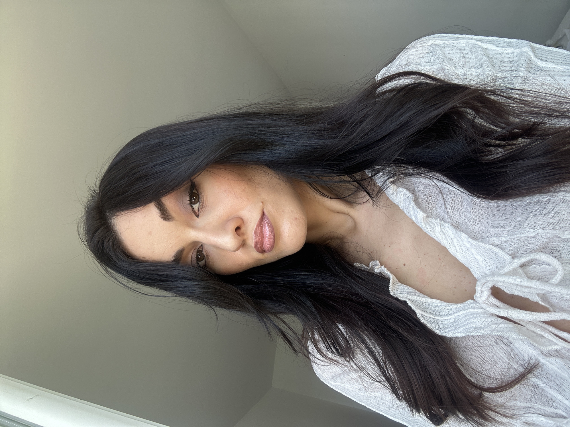 A medium shot portrait of a woman with long, dark, wavy hair. She is wearing a white, textured blouse with a V-neckline that is tied with a drawstring. The woman is looking directly at the camera with a neutral expression. The background is plain and appears to be a wall, with a window on the left side of the frame. The lighting suggests an indoor setting, possibly in a residential apartment in the city of Burgas, Bulgaria.