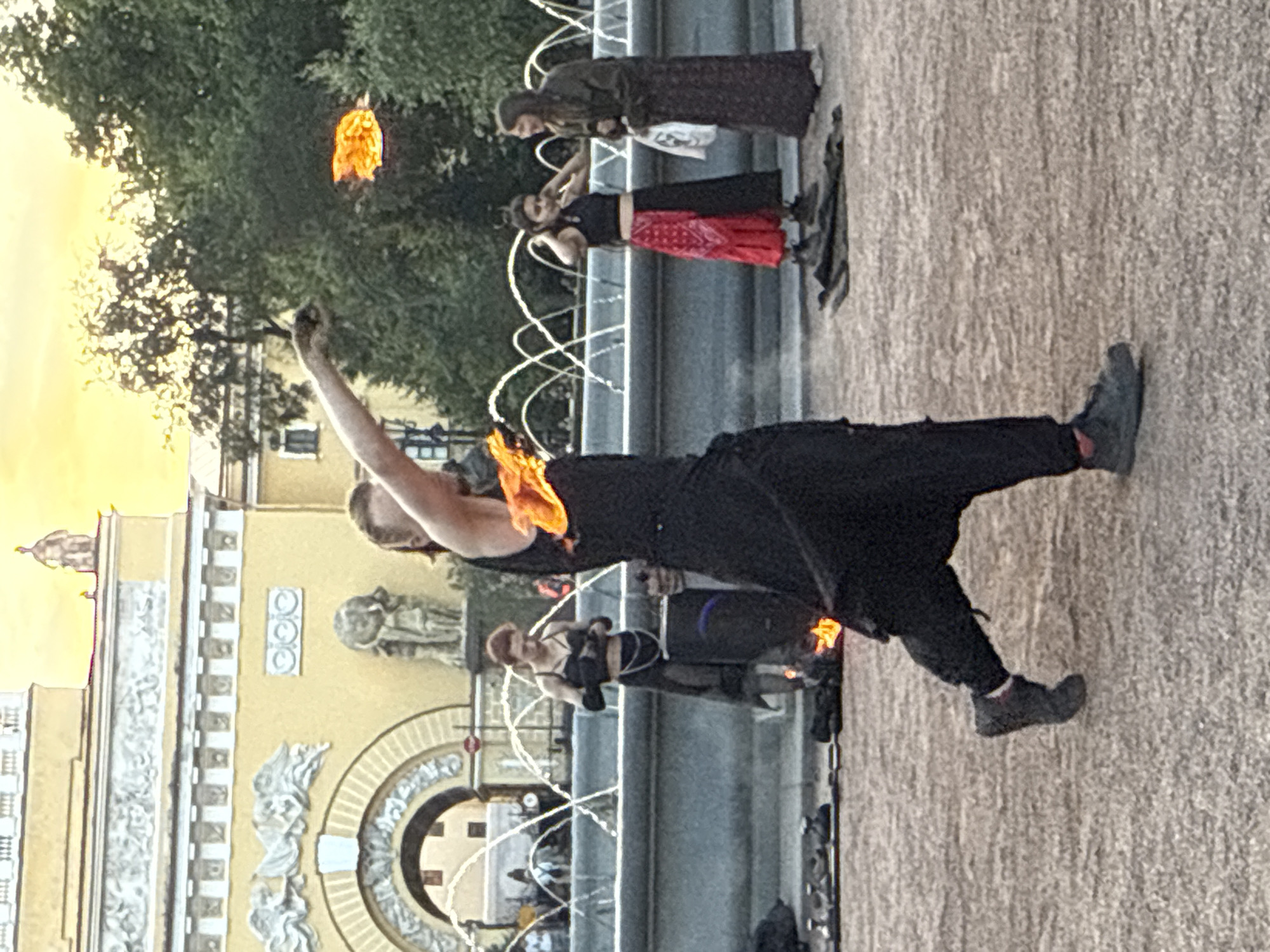 The image captures a street performance in Saint Petersburg, Russia, likely near the Admiralty building or Palace Square, as suggested by the neoclassical architecture and the general ambiance. A performer in the foreground, dressed in dark, flowing clothing, is engaged in fire dancing. Flames are visible around their torso and in their outstretched right hand. To the left of the main performer, another individual in dark attire stands near a fountain. Further to the right, two women are present; one in a black top and a red and black skirt, and the other in a long, dark dress. They appear to be observing the performance or waiting. The scene is set in an open, paved area with a fountain in the background, and trees and ornate buildings contributing to the urban environment. The sky above is a blend of orange and yellow, indicating it may be sunset or sunrise.