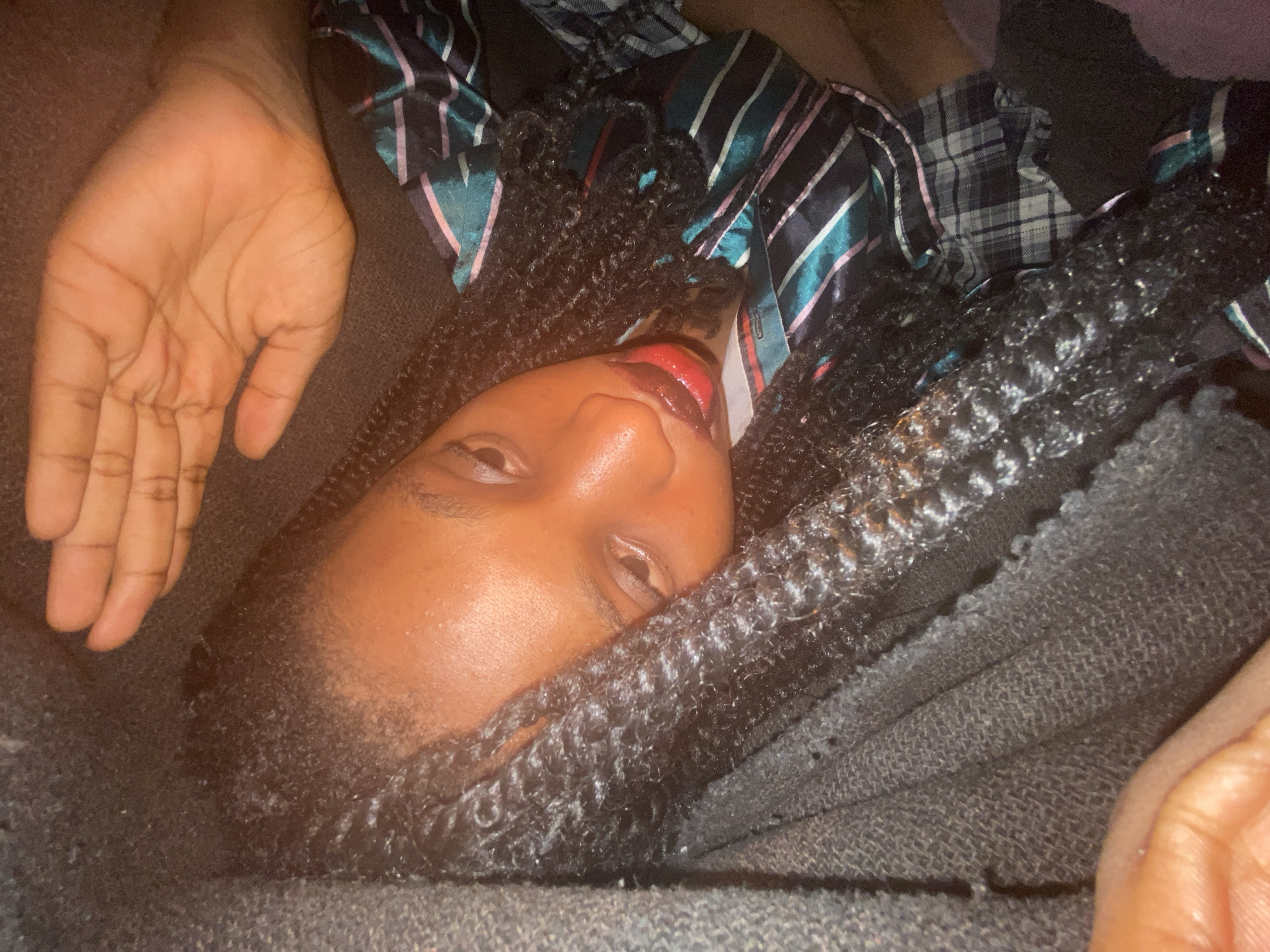 The image presents a close-up, low-angle view of a person lying down. The person has dark skin, braided hair, and is wearing red lip gloss. They are partially covered by a dark, textured blanket. Part of their face is visible, showing their eyes partially open. A hand is positioned near the upper left corner of the frame. The person is wearing a striped and plaid pajama shirt. The background is dark and out of focus. The location and city are unknown.