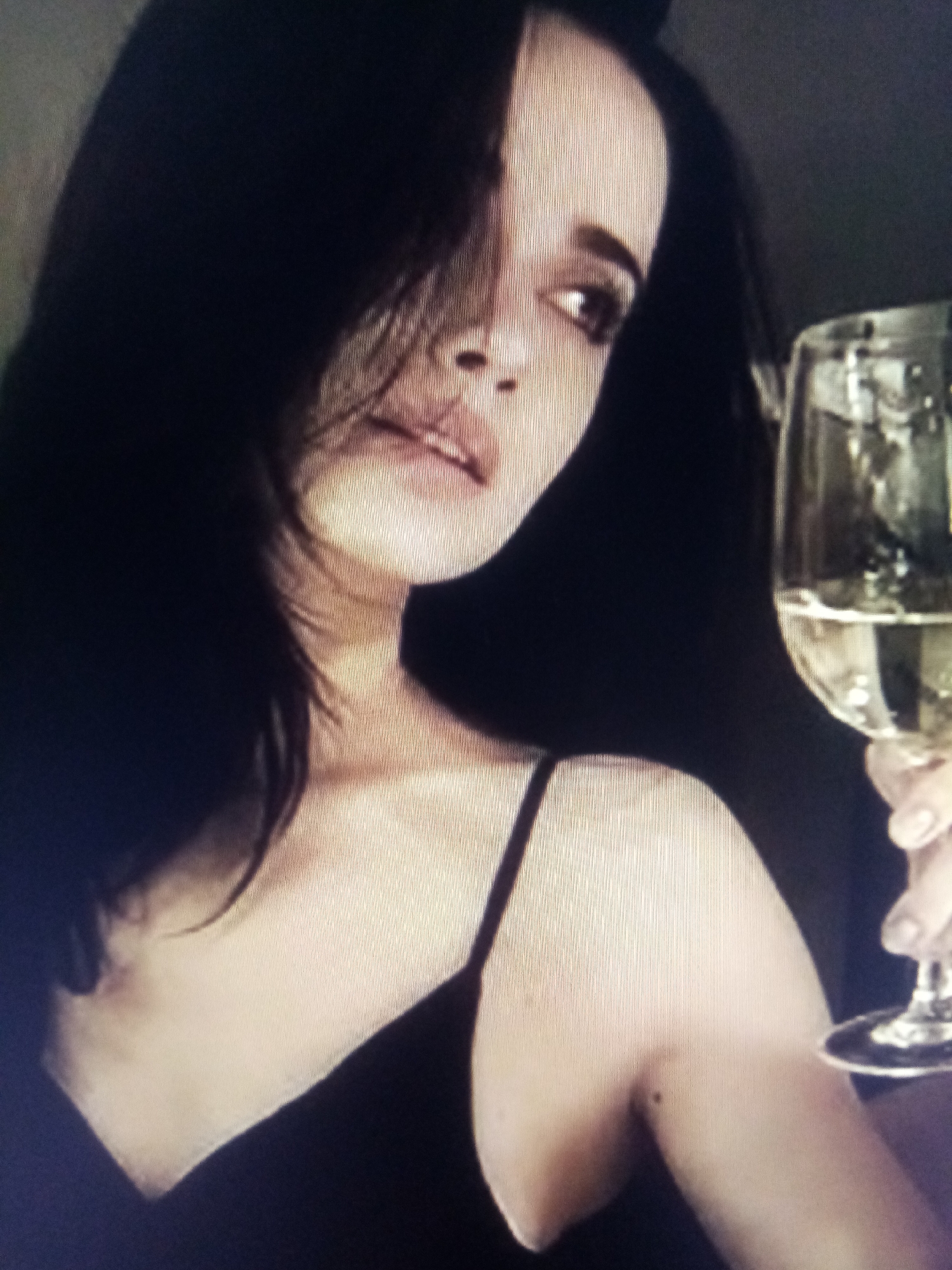 This is a medium shot of a woman with dark hair. She is wearing a black spaghetti strap top and holding a glass of white wine. Her head is turned away from the camera, and she is looking off to the side. Her hair covers part of her face. The lighting is dim, and there are some vertical lines visible in the image, suggesting it was taken from a screen. The background is dark and indistinct. The woman's expression is not clearly visible due to the angle and lighting.