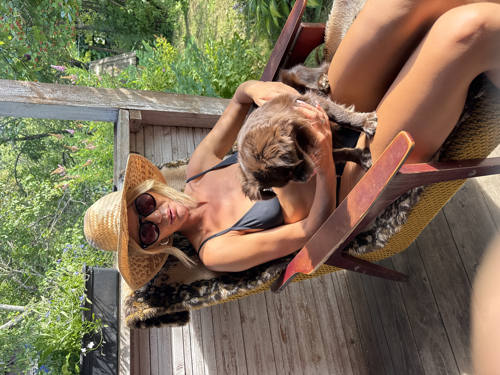 A blonde woman wearing a straw hat and sunglasses sits in a wooden chair on a wooden deck. She is wearing a black bikini and holding a small brown dog. The deck is surrounded by greenery and flowers. The scene appears to be in a garden or backyard.