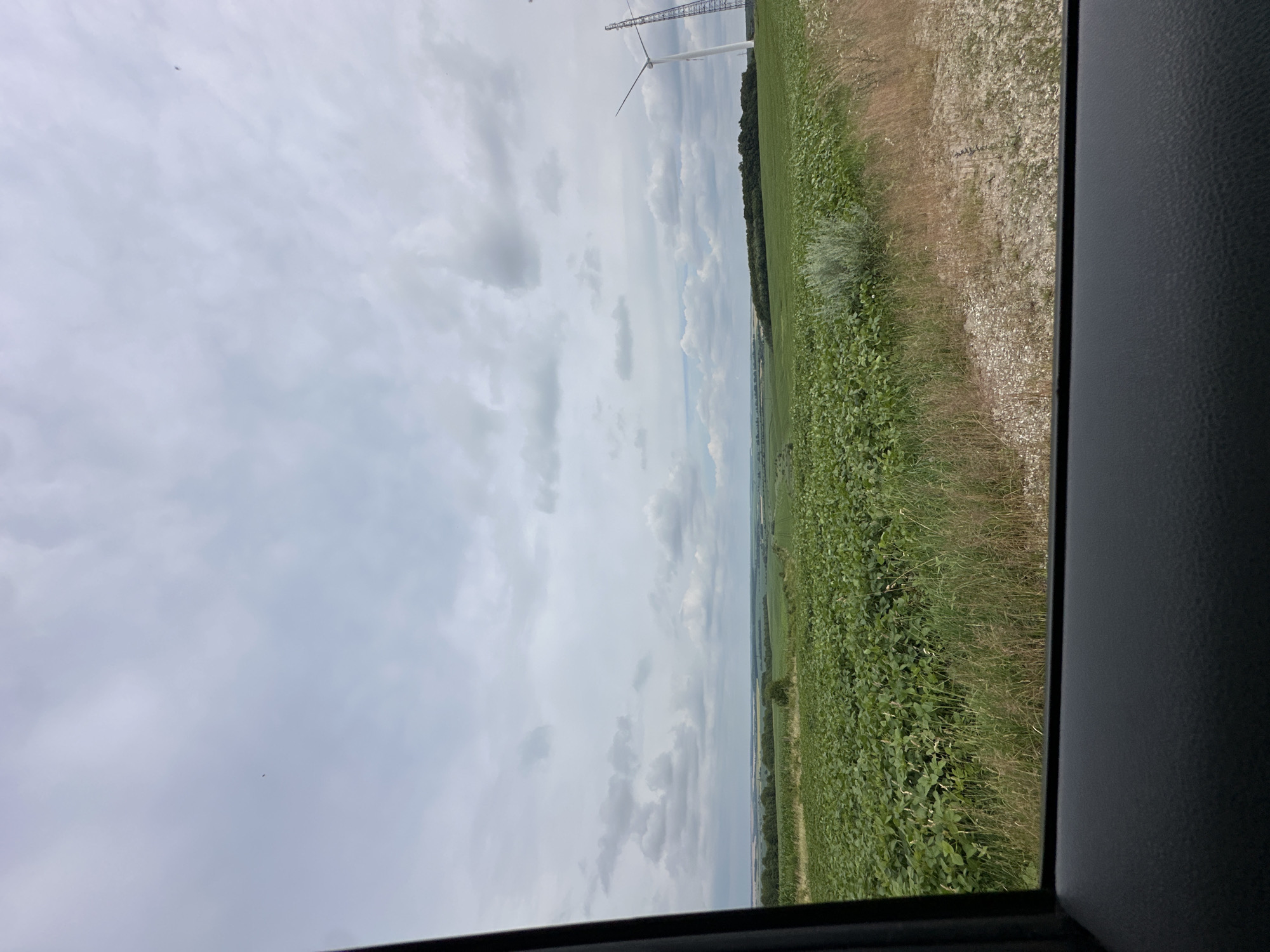 A wide shot captures a rural landscape under a cloudy sky. In the foreground, a dark car interior frames the view, suggesting the photo was taken from inside a vehicle. A vast field of lush green crops, likely soybeans, stretches across the mid-ground, bordered by dry grass and a gravelly path on the right. In the distance, rolling green hills are visible, punctuated by a line of trees. Two prominent wind turbines stand on the right side of the frame, one fully visible with its blades extended, and the other a partial view of its lattice tower. The sky is overcast with scattered clouds, casting a diffused light over the scene. The location is specified as the city of Korchivka, Ukraine. There are no people or animals visible in the image.