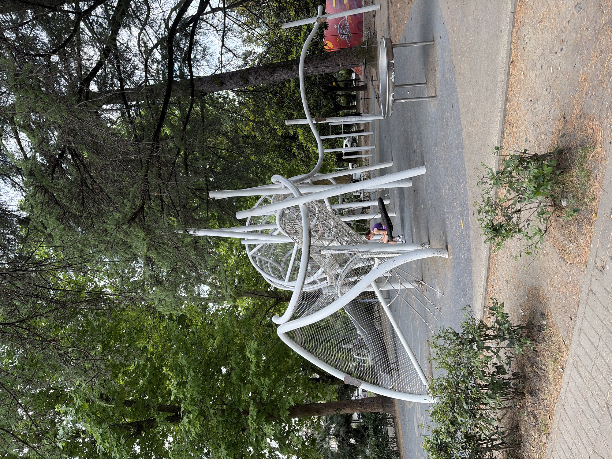 This media file captures a playground scene in Sochi, Russia. The central object is a modern, white metal climbing structure with a geodesic dome-like component and netted surfaces, designed for children's play. A person wearing a purple shirt and dark pants is crouched on the structure, possibly supervising or engaging with the equipment. The playground is situated outdoors, surrounded by lush green trees, suggesting a warm, possibly summer day with overcast skies, as indicated by the soft, diffused light. The ground surrounding the playground is a mix of paved surfaces and dry grass or fallen leaves, with some small green bushes near the edge. In the background, a portion of a red sign with white lettering is visible, though the text is not clearly legible. The overall impression is a calm, everyday moment in a public park.