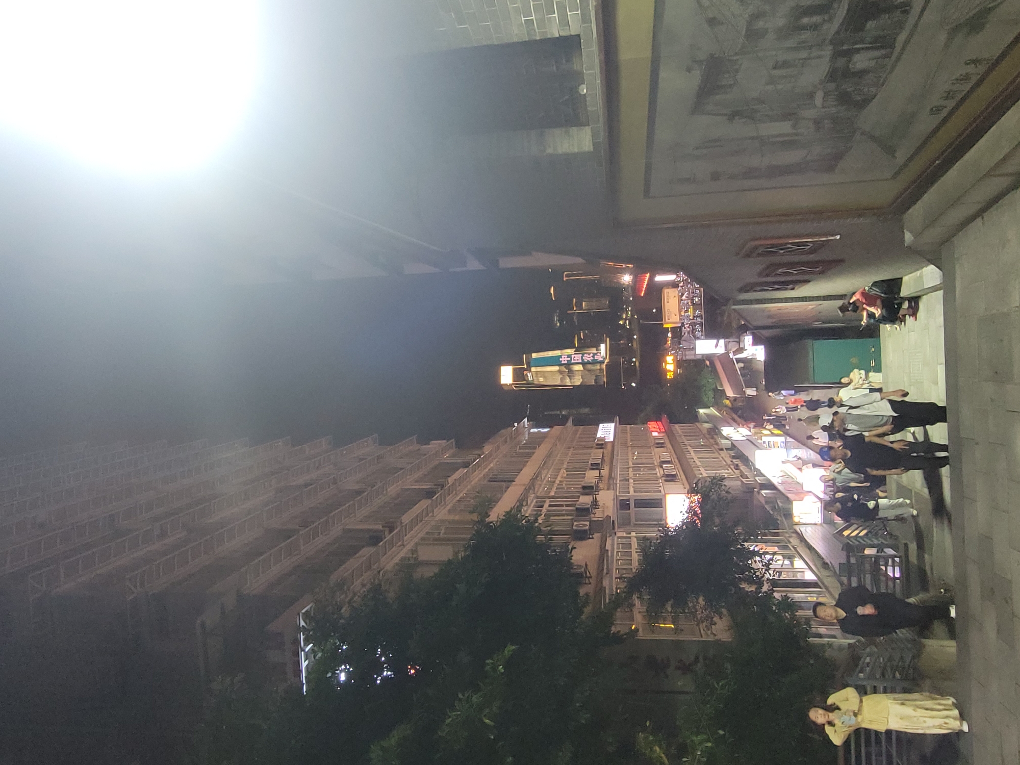 The image depicts a vibrant night scene on a bustling street in what appears to be an urban area. The overall setting suggests a commercial or residential district in Yuzhong District, China.
**Subjects:**
Several people are visible, primarily on a paved walkway to the right side of the frame. In the foreground, a woman in a light-colored dress stands, looking down. Further back, a man in dark clothing is seated on a bench. A group of people stand behind him, some wearing dark attire, one in a white shirt, and another with a blue cap, seemingly gathered or queuing.
On the left side of the street, a tall, multi-story building with numerous balconies and air conditioning units dominates the mid-ground. Further back and slightly left of center, a prominent skyscraper with an illuminated sign is visible. Closer to the right-hand walkway, there are street-level shops with bright signs. Trees and bushes are interspersed along the street, adding greenery to the urban landscape.
**Activity or event taking place:**
People are walking, standing, and possibly socializing or dining outdoors. The illuminated shopfronts suggest an active commercial area where people are engaged in evening leisure or errands.
**Notable details:**
The scene is set at **night**, illuminated by abundant artificial lights from buildings, street lamps, and shop signs, creating a lively atmosphere. A very bright light source or lens flare is present in the upper-left area of the frame. On an overhead wall above the people on the right, a large **mural or artwork** depicting an older architectural scene or map adds a historical or cultural element. The architecture and illuminated signs are characteristic of a Chinese city.
**Visible Text:**
The most clearly discernible text is "中国银行" (Zhōngguó Yínháng), which translates to "Bank of China," displayed prominently on the illuminated sign of the distant skyscraper. Other shop signs bear Chinese characters but are too distant or blurry to be read distinctly.