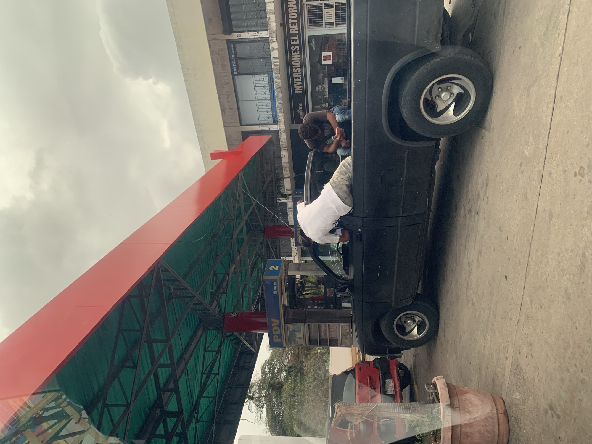 The image displays an exterior scene at a PDVSA gas station in La Fuente, Venezuela, under an overcast sky. In the foreground, a black pickup truck is positioned, containing two males in its open bed. The male on the left is observed from the rear, wearing a white short-sleeved shirt and light-colored patterned shorts, leaning forward as if reaching into the truck's cab or examining something below. The male on the right, clad in a dark short-sleeved shirt and blue jeans, is seated, engrossed in a small, light-colored handheld device. The black pickup truck features silver five-spoke wheels. Behind and to the left of the pickup, a red hatchback car is partially visible, bearing a Venezuelan license plate "AD2090S". The gas station structure includes a red upper canopy over a green metal truss system. A blue and yellow gas pump unit, marked with "PDV" and "PDVSA" logos and the number "2", stands beneath the canopy. A red pillar nearby also displays the number "3". In the background, a multi-story building hosts various businesses, with visible signs reading "INVERSIONES EL RETORNO," "BIG PC.CA," and "Santa Teresa." The ground surface is concrete. A terracotta potted plant is partially visible in the bottom-left corner, viewed through a reflective surface.