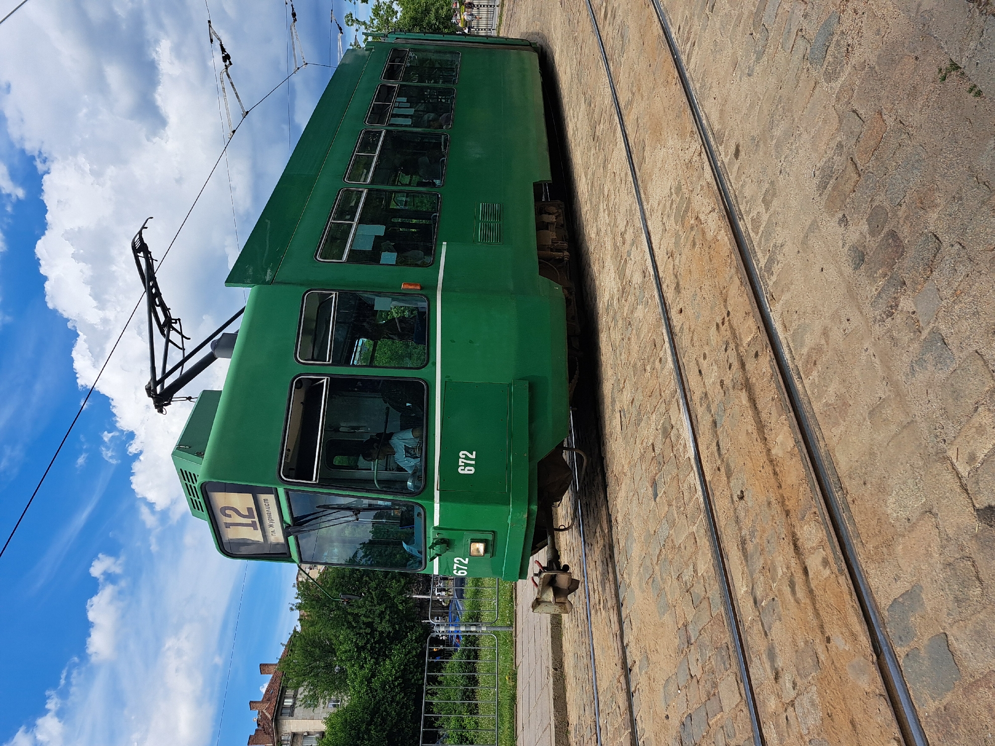 A bright green tram, numbered 672, is stopped on a cobblestone street, indicating a city setting. The tram has a route sign displaying "12" and "ул. Журналист". A person, presumably the driver, is visible through the front window. The tram's pantograph is connected to overhead wires. In the background, buildings and trees are present. The sky is blue with scattered clouds.
