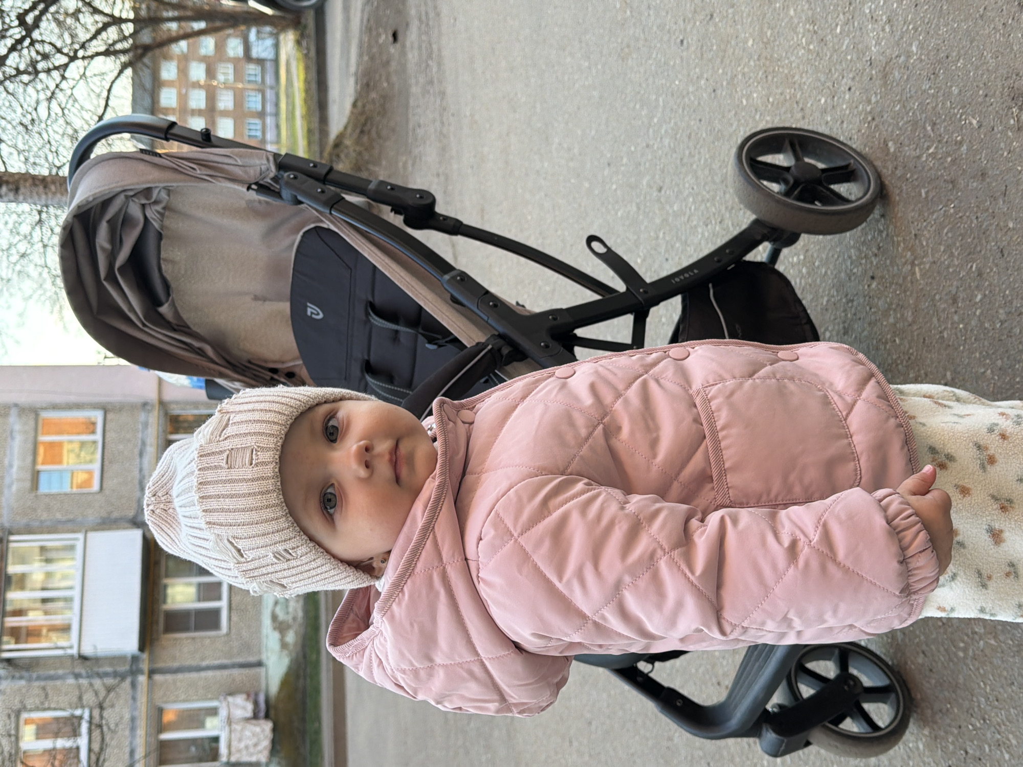 A baby in a pink quilted jacket and a white knit hat is seated in a stroller on a paved surface. The stroller has a black frame and beige fabric, visible at the top. The background includes a multi-story apartment building with visible windows and some trees. The scene appears to be outside, possibly in a residential area. This image was taken in Reshetnikovo, Russia.