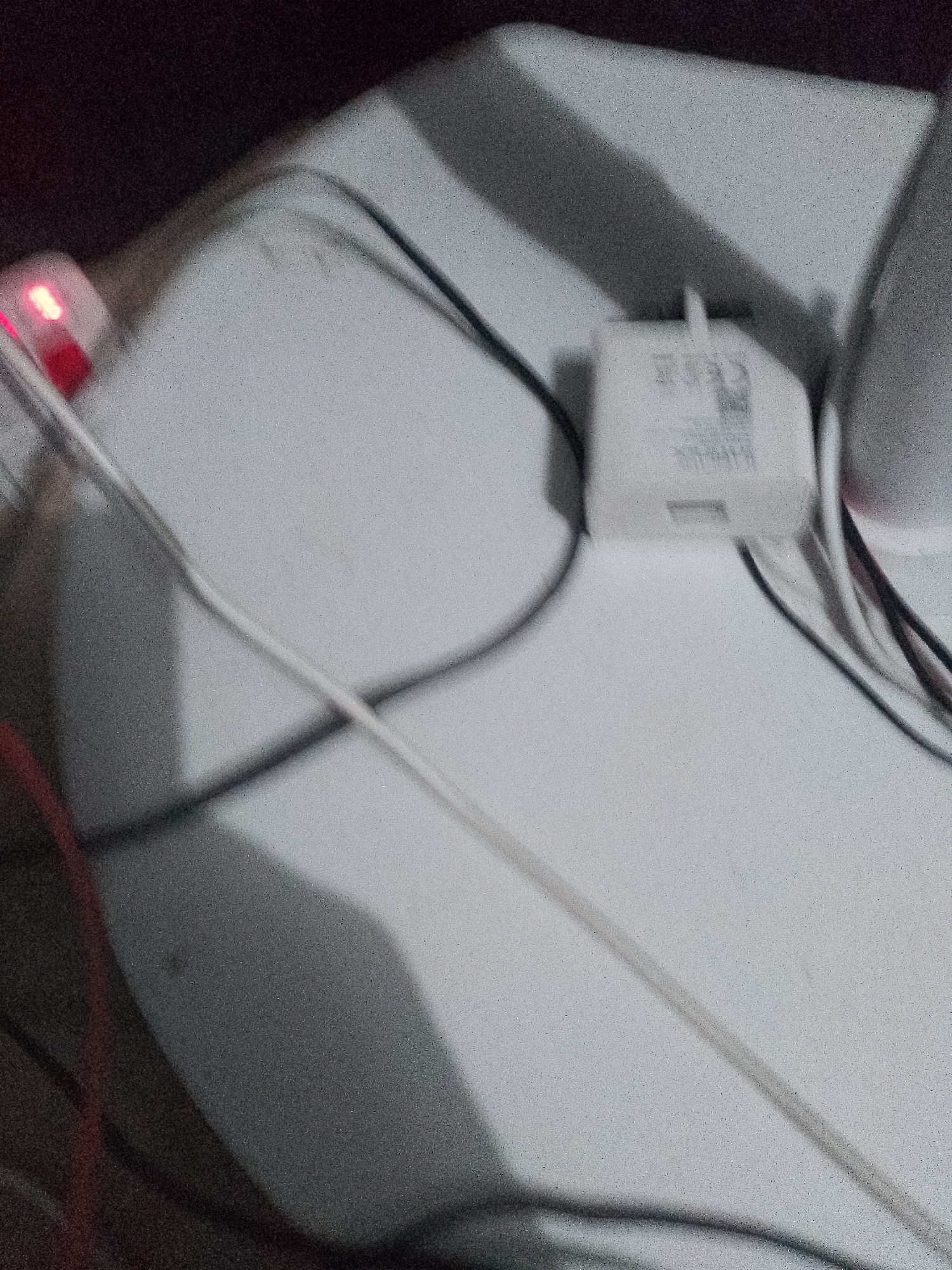 The image shows a close-up of a white power adapter with black and white wires connected to it. A red LED light is visible on a small rectangular object to the left of the adapter. The background is blurry and appears to be a textured surface. The scene is likely indoors. There is no indication of a specific location in Jauro Yinu, Nigeria.