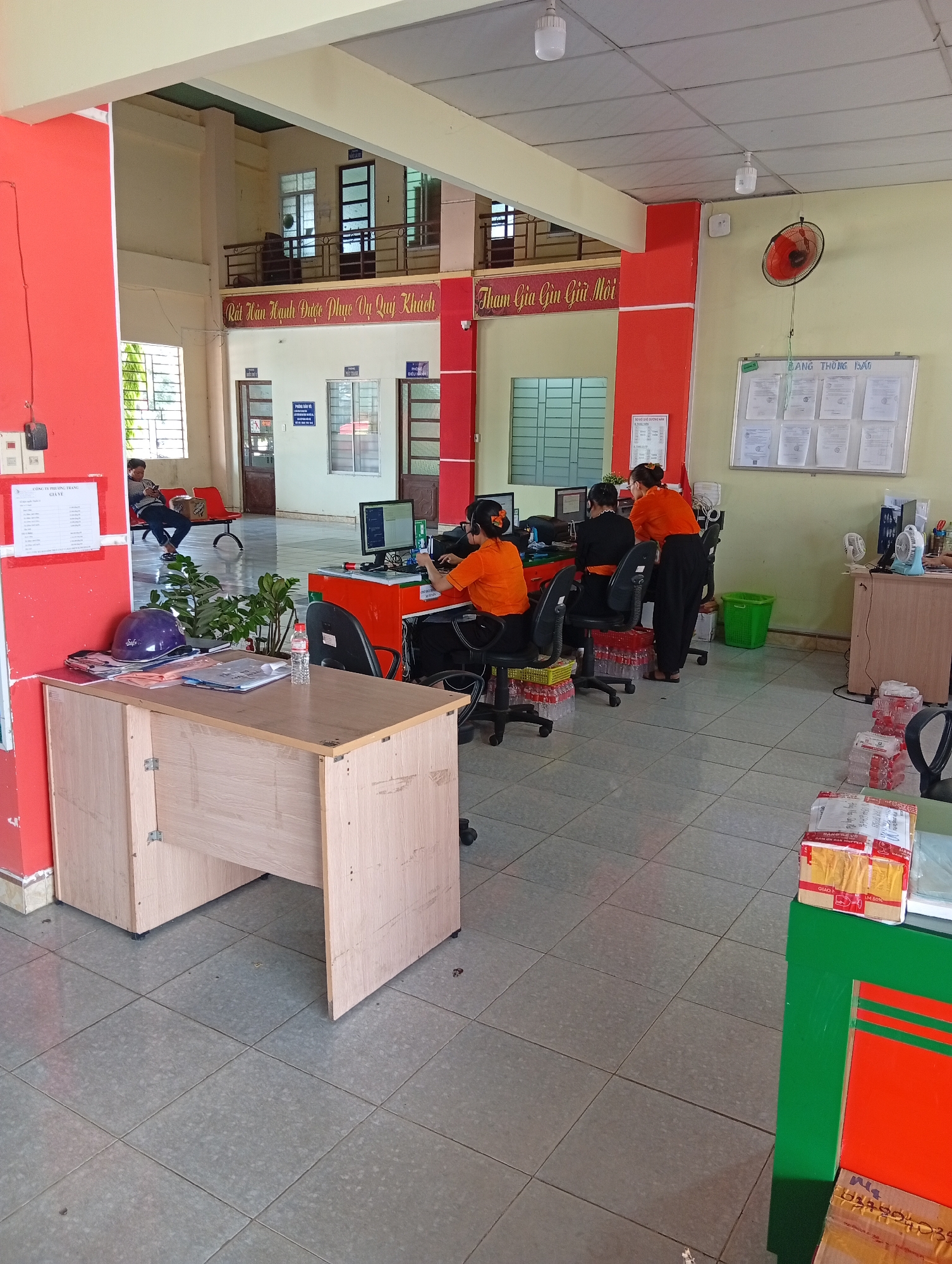 The image depicts an interior scene in what appears to be an office or service area, potentially located in Phan Rang – Tháp Chàm, Vietnam. Three women wearing orange shirts are seated at computer workstations. One woman is standing, interacting with the seated women. A table and a desk with office items are in the foreground. Signage in Vietnamese is present on the walls. In the background, a man is seated on a bench. The environment is a tiled floor and the room has a functional, public service feel. Packages are also visible on the floor.