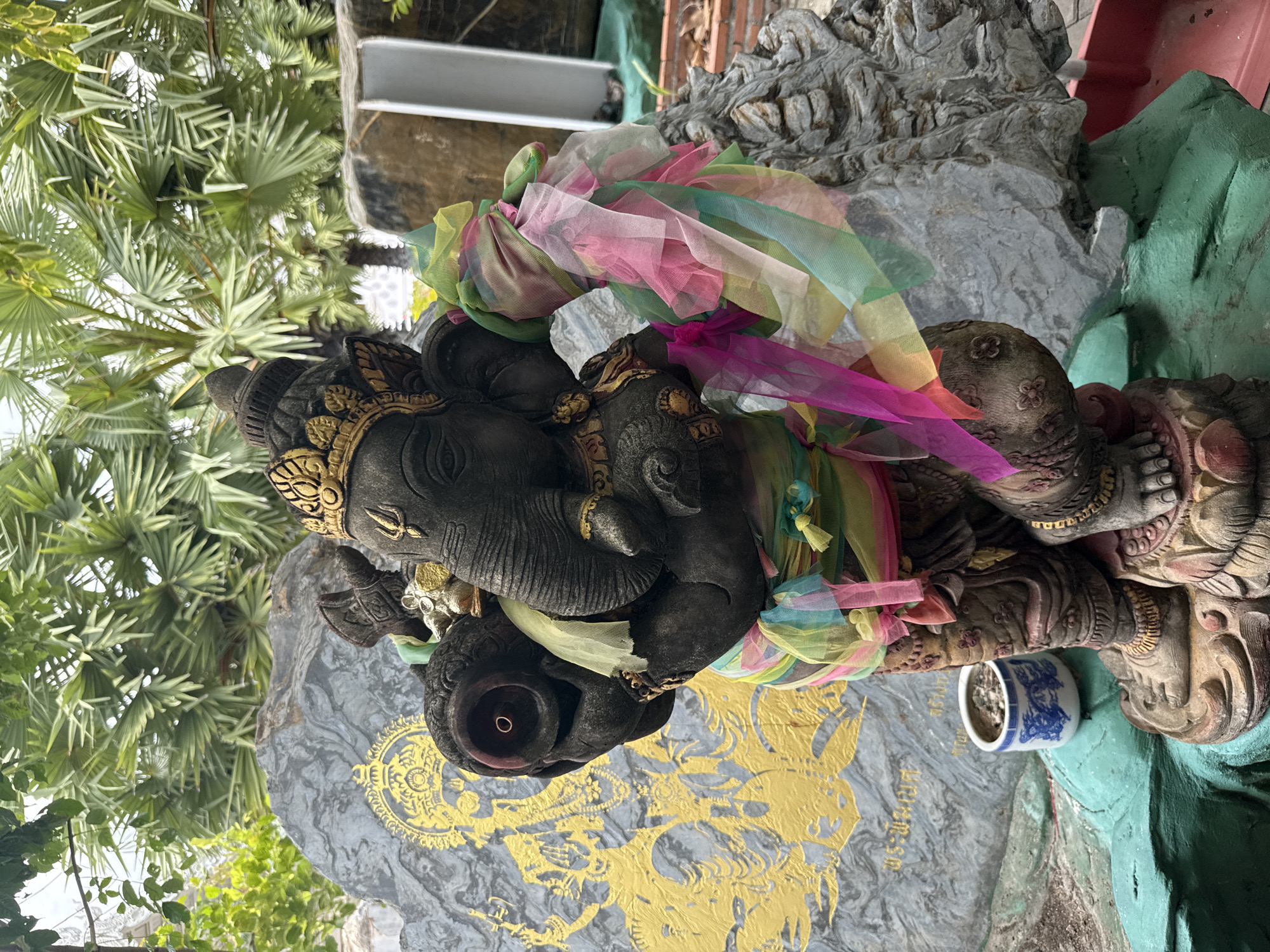 A weathered stone statue of Ganesha stands in an outdoor setting with palm trees in the background. The statue is dark gray, with some gold accents on its headdress and jewelry. It is adorned with colorful, sheer fabric strips tied around its torso and arms. To the left of the statue is a large, gray rock with a golden depiction of a deity. In front of the statue, to the left, is a small, blue and white ceramic pot containing ash. The ground around the statue is painted green, and some reddish-brown bricks are visible in the lower right corner of the image. The scene appears to be in Tambon Na Chom Thian, Thailand.