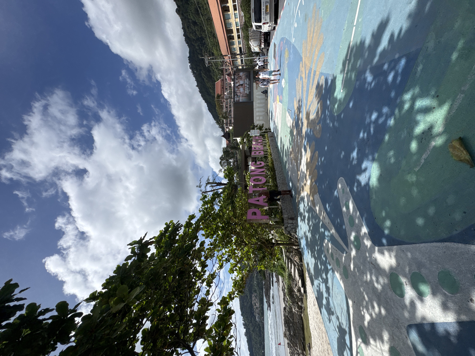 The image captures a sunlit day in Patong, Thailand, with a vibrant blue sky dotted with white clouds. In the foreground, a colorful, painted pathway with abstract designs in blue, green, and yellow shades leads the eye toward the left. Shadows of tree leaves are cast across this path, creating dappled patterns. To the left, lush green trees with broad leaves overhang the scene, partially obscuring the sky.
Behind the pathway, a sandy beach meets rocky shorelines and the gentle waves of the ocean. A red flag is visible near the water's edge. A prominent pink sign displaying "PATONG BEACH" in large letters stands near the pathway, with a person in dark shorts and a dark shirt standing behind it.
Further in the background, a row of buildings, typical of a tourist area, lines the coast. These buildings have terracotta-tiled roofs and light-colored facades, with balconies and windows visible. One building features a large billboard with an advertisement. A white van and a truck are parked on a road adjacent to the buildings. Several people are walking along the pathway and the road in the middle ground. The overall scene depicts a lively coastal area in Patong, Thailand, on a bright, clear day.