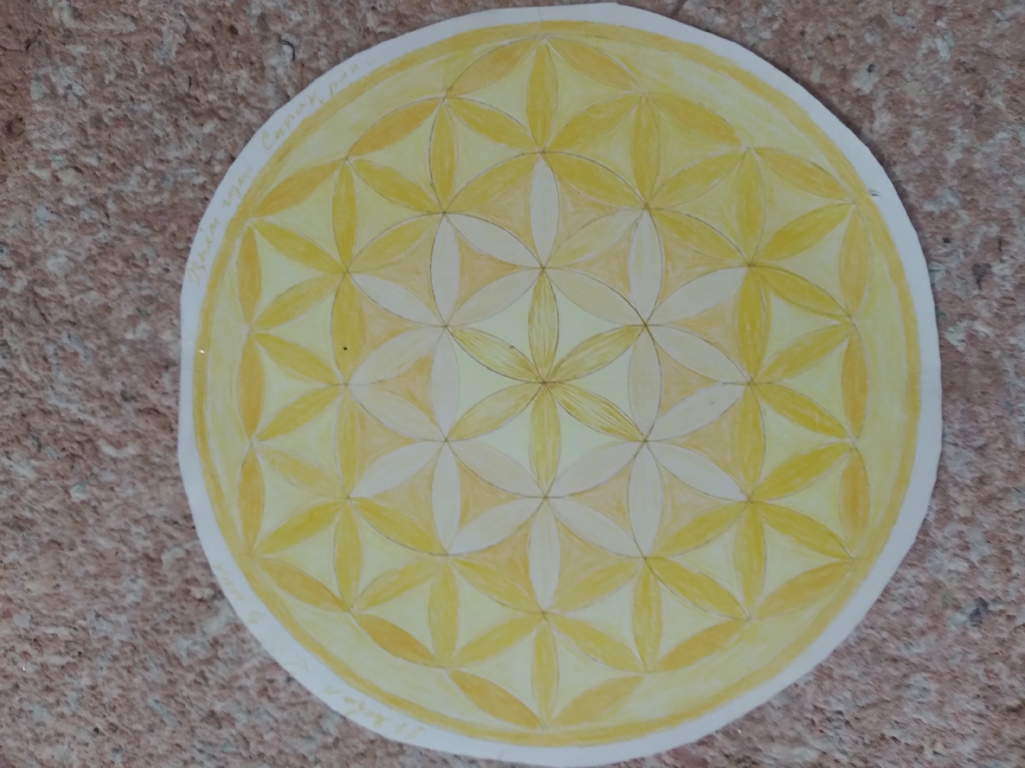 The image shows a yellow and orange watercolor drawing of the Flower of Life geometric pattern on a white circular piece of paper. The drawing is placed on a textured, reddish-brown surface, possibly a floor or wall. There are faint handwritten words in a light color on the outer edge of the drawing, on the right side. The location is identified as Baki, Uzbekistan.