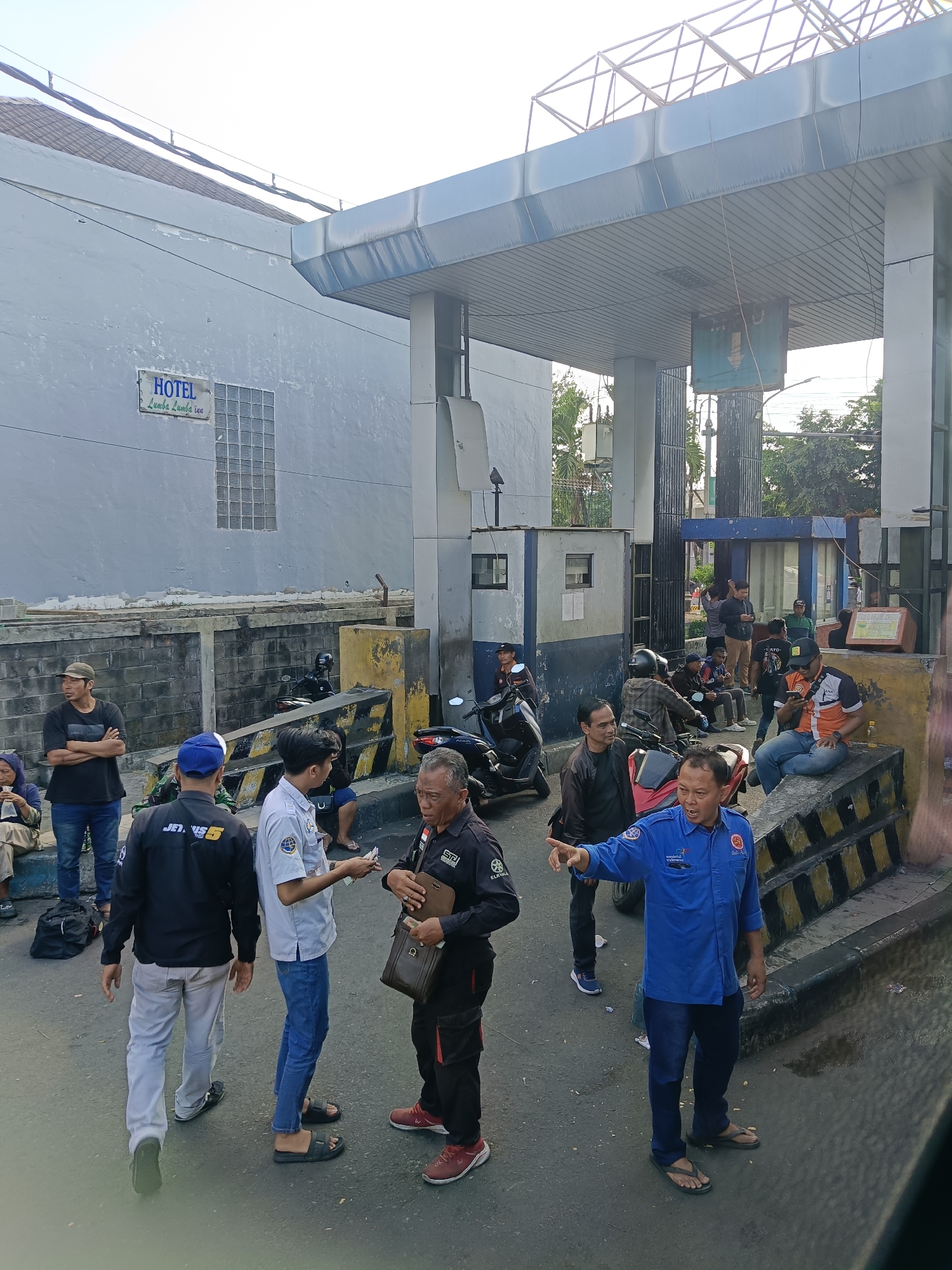 The image depicts a street scene in Kedungrejo, Indonesia, near what appears to be a toll gate or a similar checkpoint. The environment includes a building with a sign that reads "HOTEL Lumba Lumba Inn." Several individuals are present, some standing, others seated on a concrete barrier. A group of men are engaged in conversations, with some wearing uniforms that suggest they may be security personnel or employees of a service. One man in a blue shirt with badges is actively gesturing and speaking, appearing to direct or instruct others. Motorcycles are parked in the background, indicating transportation methods in the area. The overall scene suggests a functional space where people are gathering or passing through, with elements of commerce and public service visible. The architecture includes a covered structure with white columns and a metallic roof, typical of infrastructure found in such locations. The ground is paved with asphalt, and there are yellow and black striped barriers in the foreground. The lighting suggests it is daytime, with overhead wires and trees visible in the background.