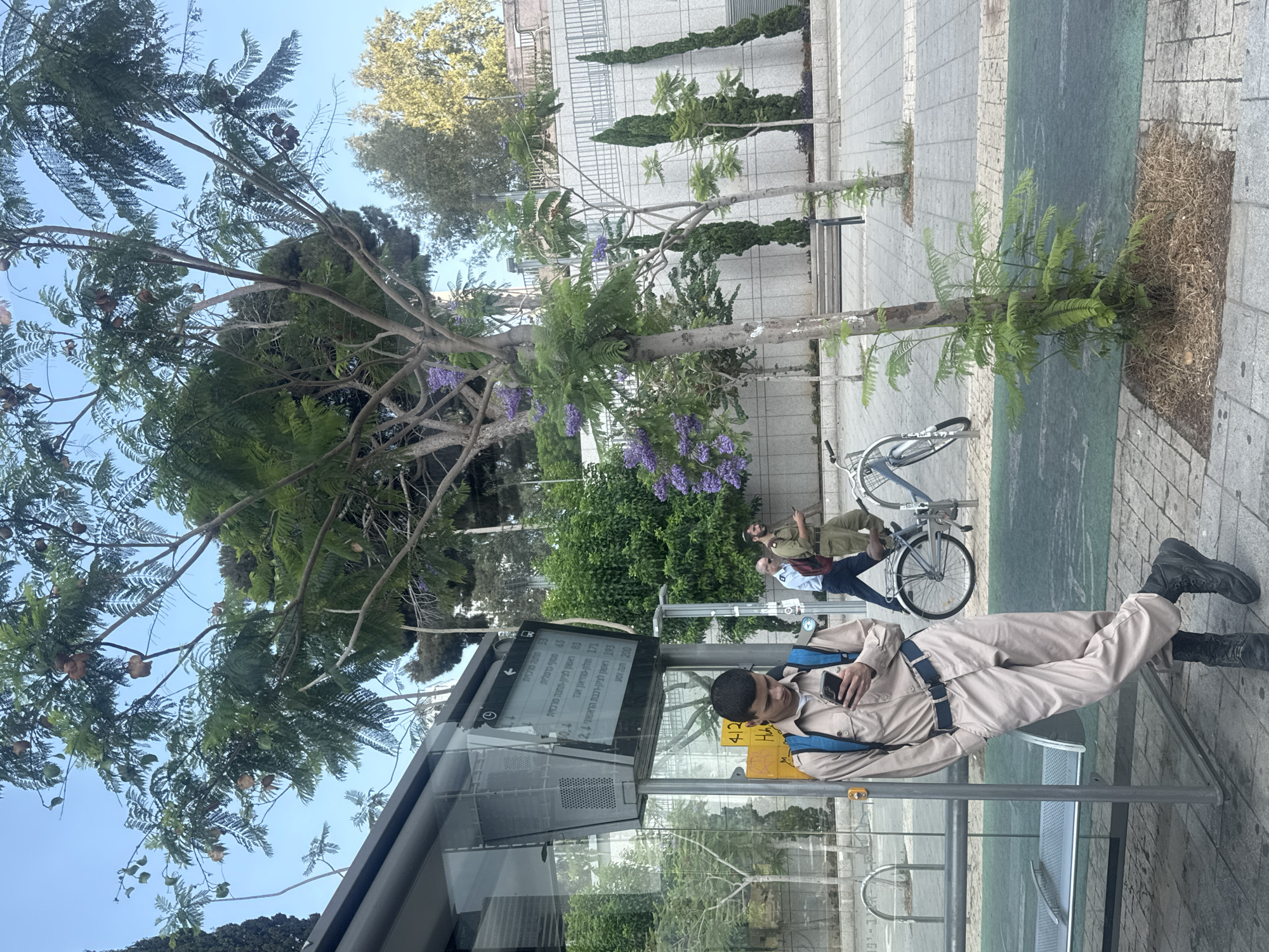 A young man in a tan uniform and blue backpack, wearing black boots, stands at a bus stop in Tel Aviv, Israel, looking at his phone. He leans against a glass and metal bus stop shelter. The bus stop has a digital display showing bus routes and times. In the background, two men in military uniforms walk past. A bicycle is parked near them. The scene is set against a backdrop of trees, some with purple flowers, and a light-colored tiled wall. The ground is paved with interlocking concrete tiles and a green painted stripe.