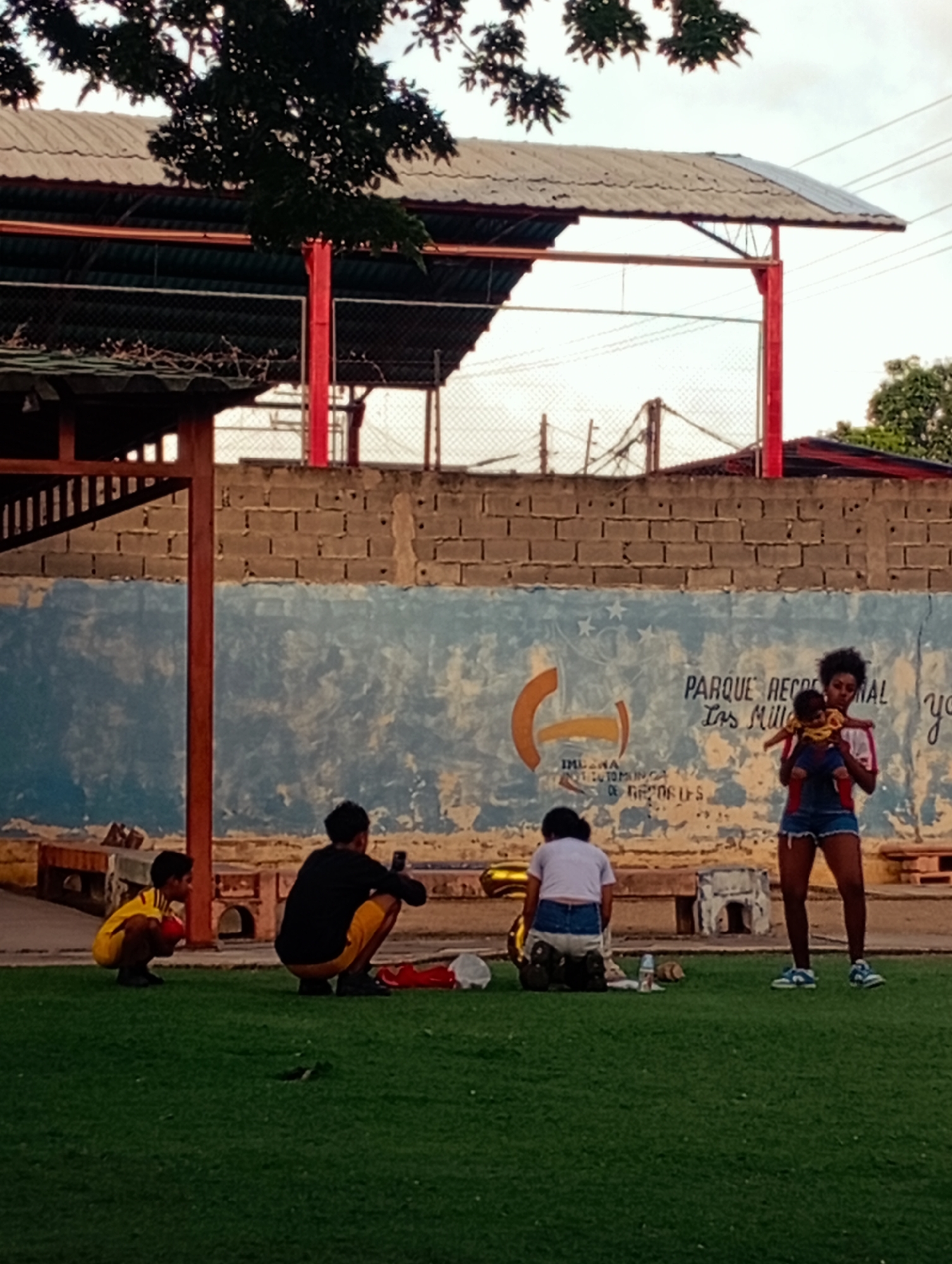 In Juan Griego, Venezuela, on a grassy area with a brick wall backdrop, four individuals are present. A young boy wearing a yellow shirt and shorts crouches on the left, holding a red ball. Next to him, a person in a black shirt and yellow shorts squats while looking at a phone. In the center, another person in a white shirt and denim shorts kneels, with items scattered around them, including a shiny gold balloon. On the right, a woman with dark, curly hair, wearing a red shirt and denim shorts, stands and holds a baby in her arms. The brick wall features a mural with Spanish text and graphics, including "PARQUE RECREACIONAL" and "las Milla". The scene appears to be an outdoor recreational space.