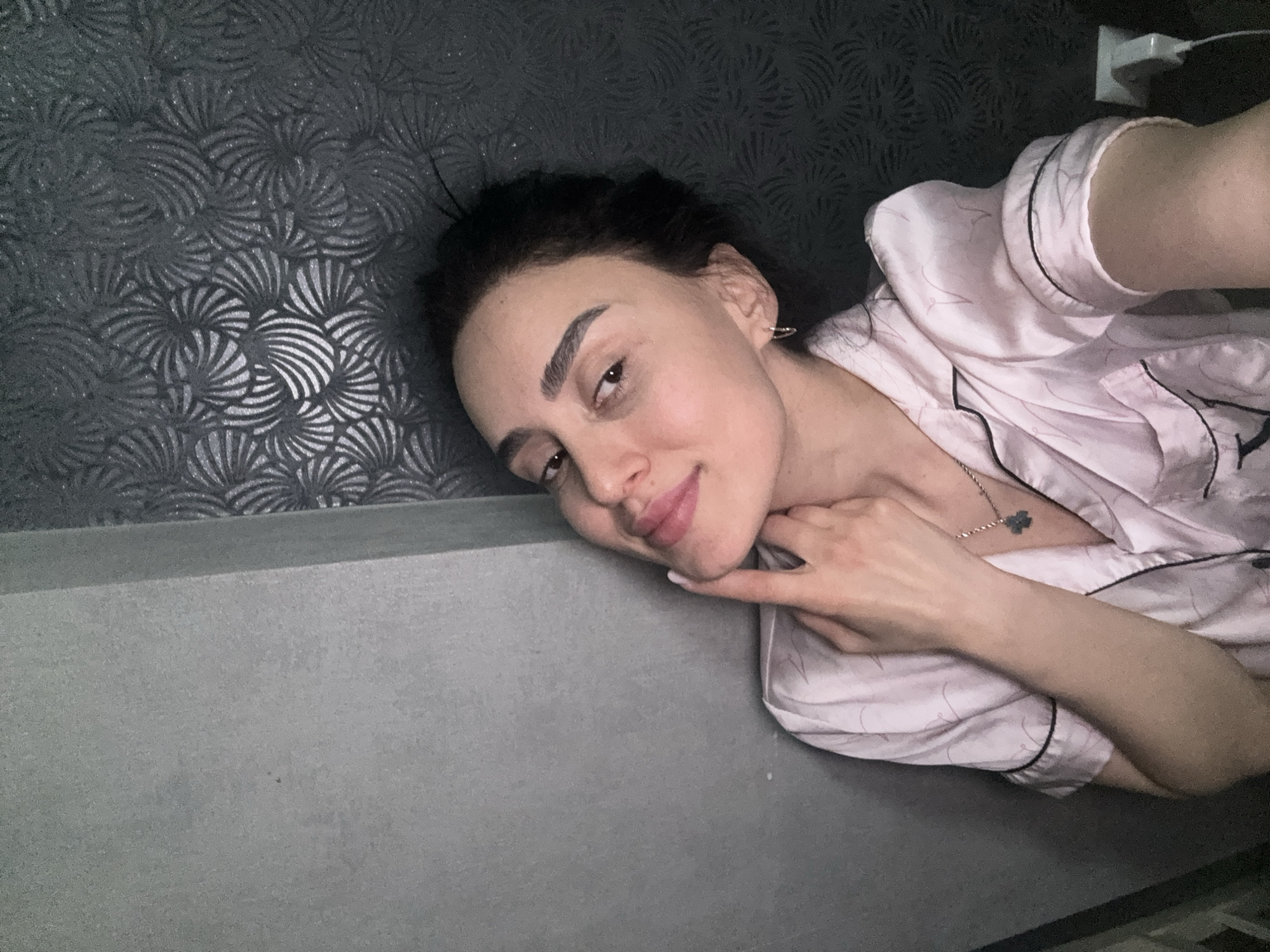 A woman is lying on a gray surface, possibly a bed or floor, with a gray patterned wall behind her. She is wearing pink pajamas with black piping and a clover-shaped pendant necklace. Her dark hair is pulled back, and she is lightly smiling, resting her chin on her hand. A charger is partially visible in the upper right corner of the image. The location is not identifiable as Tbilisi, Georgia, based on visual information.