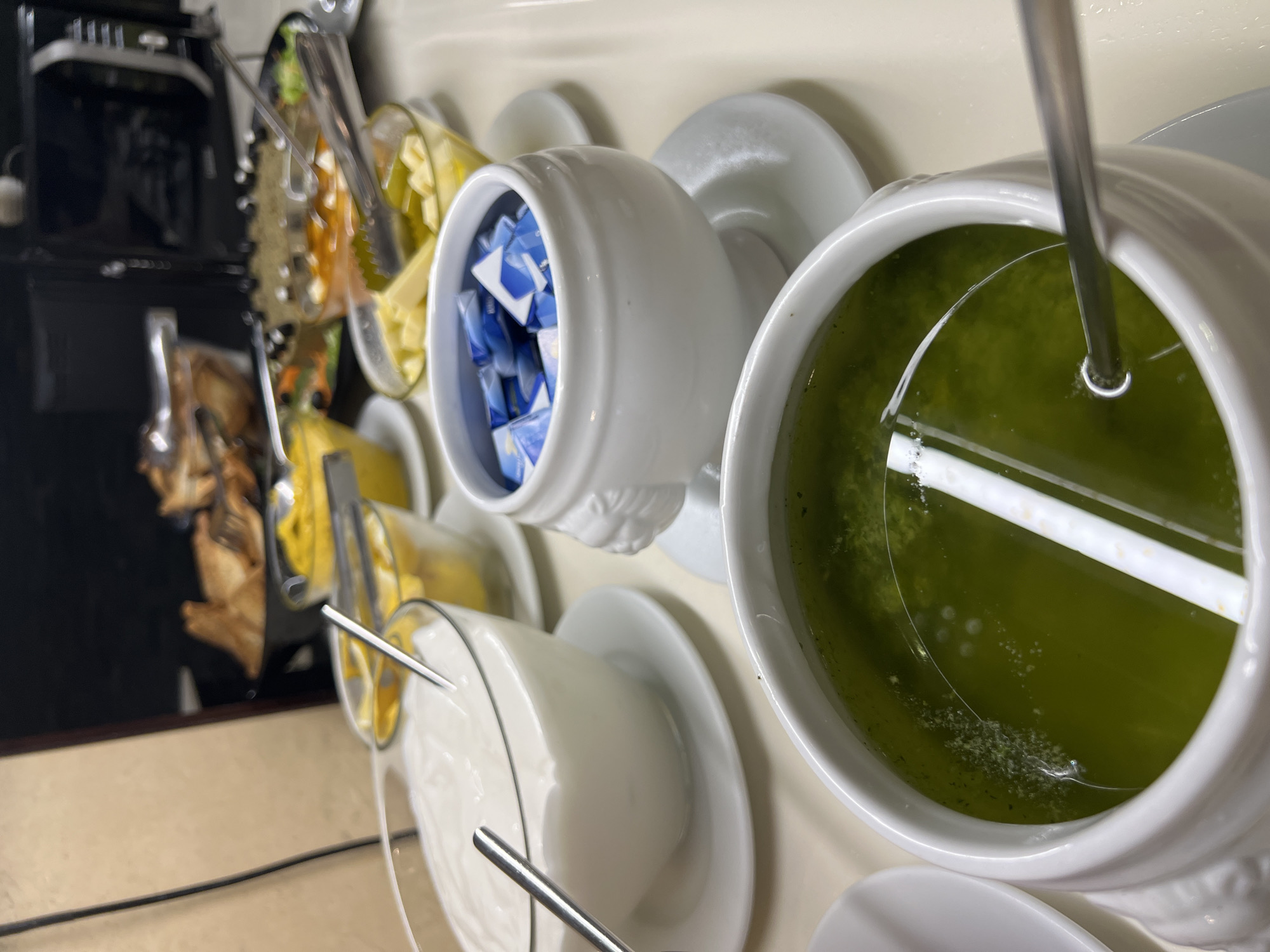 The image shows a breakfast buffet at the Hotel Riva in Petrovac centar, Montenegro. A line of white ceramic containers sits on a light-colored countertop. The containers hold: a green liquid (possibly pesto or a sauce), sugar packets in blue wrappers, yellow cheese cubes, and a white creamy substance (possibly yogurt or sour cream). Silver serving utensils are placed in each container. In the background, a black coffee machine and other food items are visible.