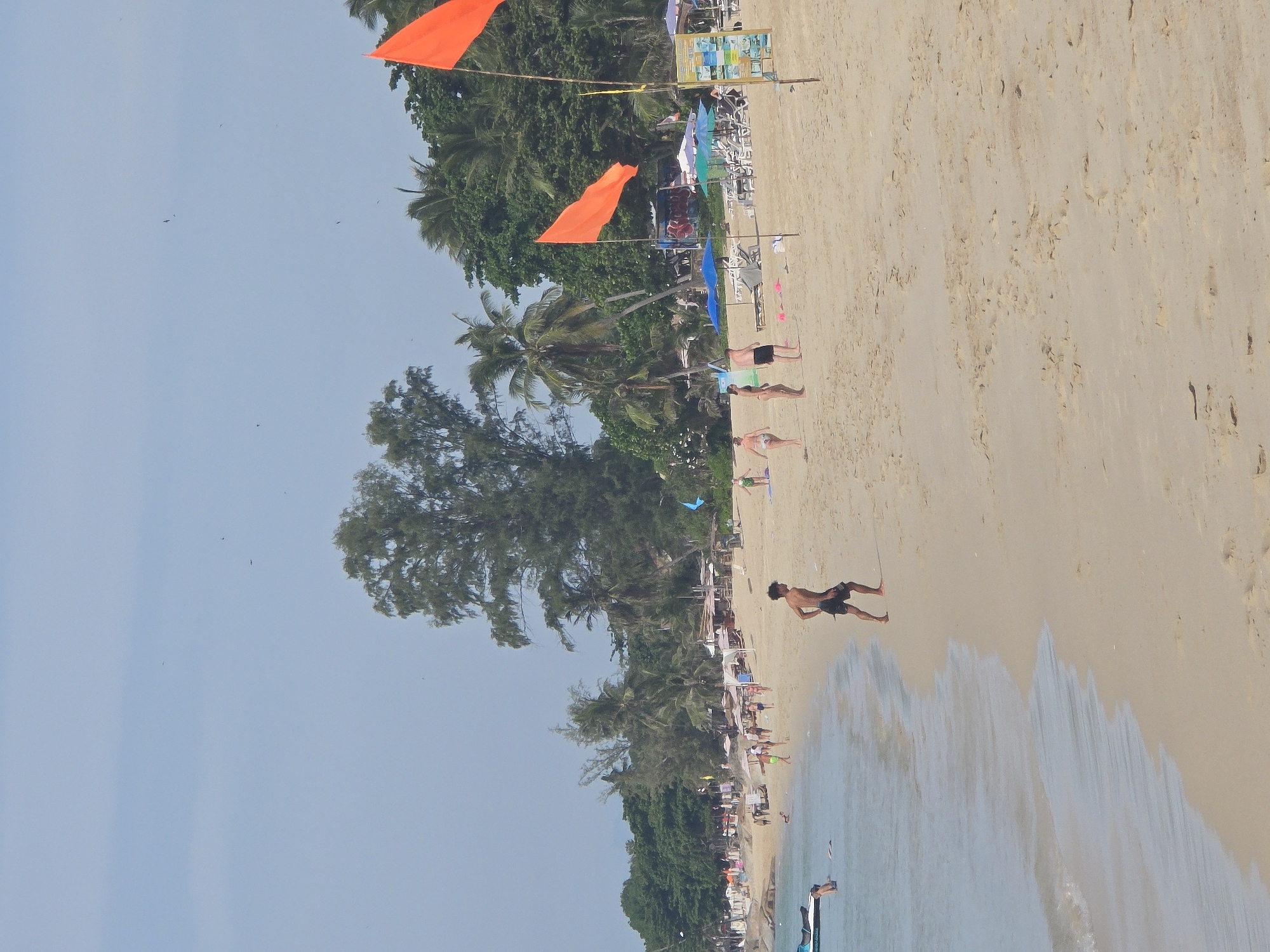 The image shows a wide, sandy beach with several people enjoying a sunny day. In the foreground, a young man in black shorts is walking on the sand, his body turned to the left. To his left, a group of people are gathered near the water's edge, some wading in the shallow waves. Further down the beach, a few individuals are walking or standing, and a jet ski is partially visible in the water on the far left. In the background, the beach is lined with palm trees and other vegetation, with a few small buildings or stalls visible. Two bright orange flags are prominently displayed on poles near the center of the frame. The sky is a clear, pale blue. The overall scene depicts a tropical beach setting with recreational activities.