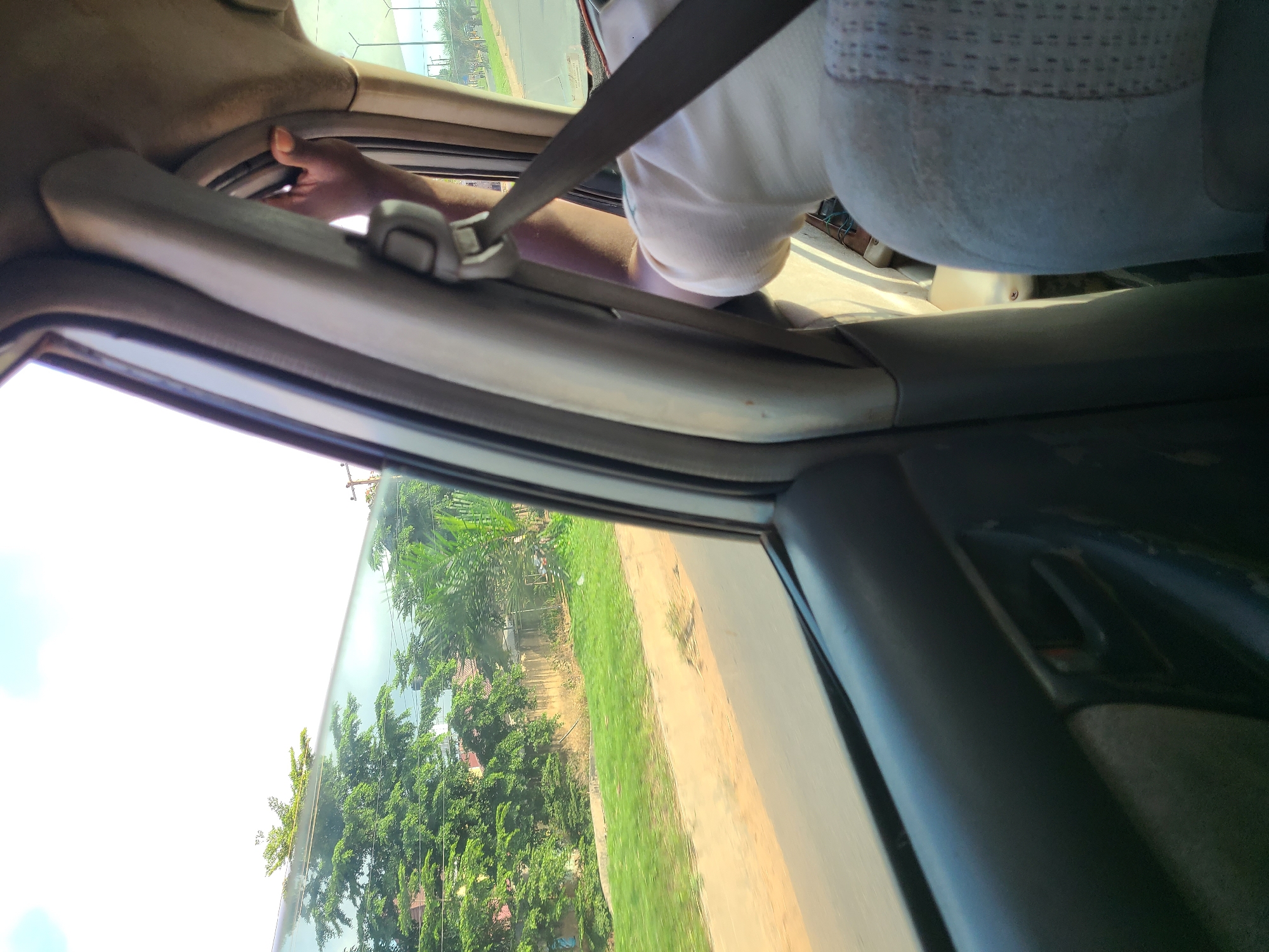 The image is taken from inside a vehicle traveling on a road in Karu, Nigeria. The view is through the passenger-side window, looking outwards and slightly upwards. Lush green trees and vegetation line the roadside, with buildings visible in the background. A hand, presumably of a passenger, is visible near the interior of the car, resting on the pillar and holding the seatbelt. The interior of the car, including a seatbelt, a door panel, and part of a seat, are also visible. The sky is bright and appears to be daytime.