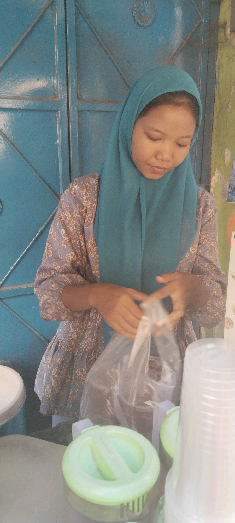 A young woman wearing a teal hijab and a floral print dress is standing and preparing a plastic bag in front of a blue metal door. Her hands are clasped together, holding the opening of the bag. To her right, clear plastic cups are stacked. In the foreground, a green lidded container is visible. The scene appears to be outdoors, possibly at a market stall, given the arrangement of the items and the background of a blue, weathered door with some peeling paint on the right. The setting is in the city of West Jakarta, Indonesia.