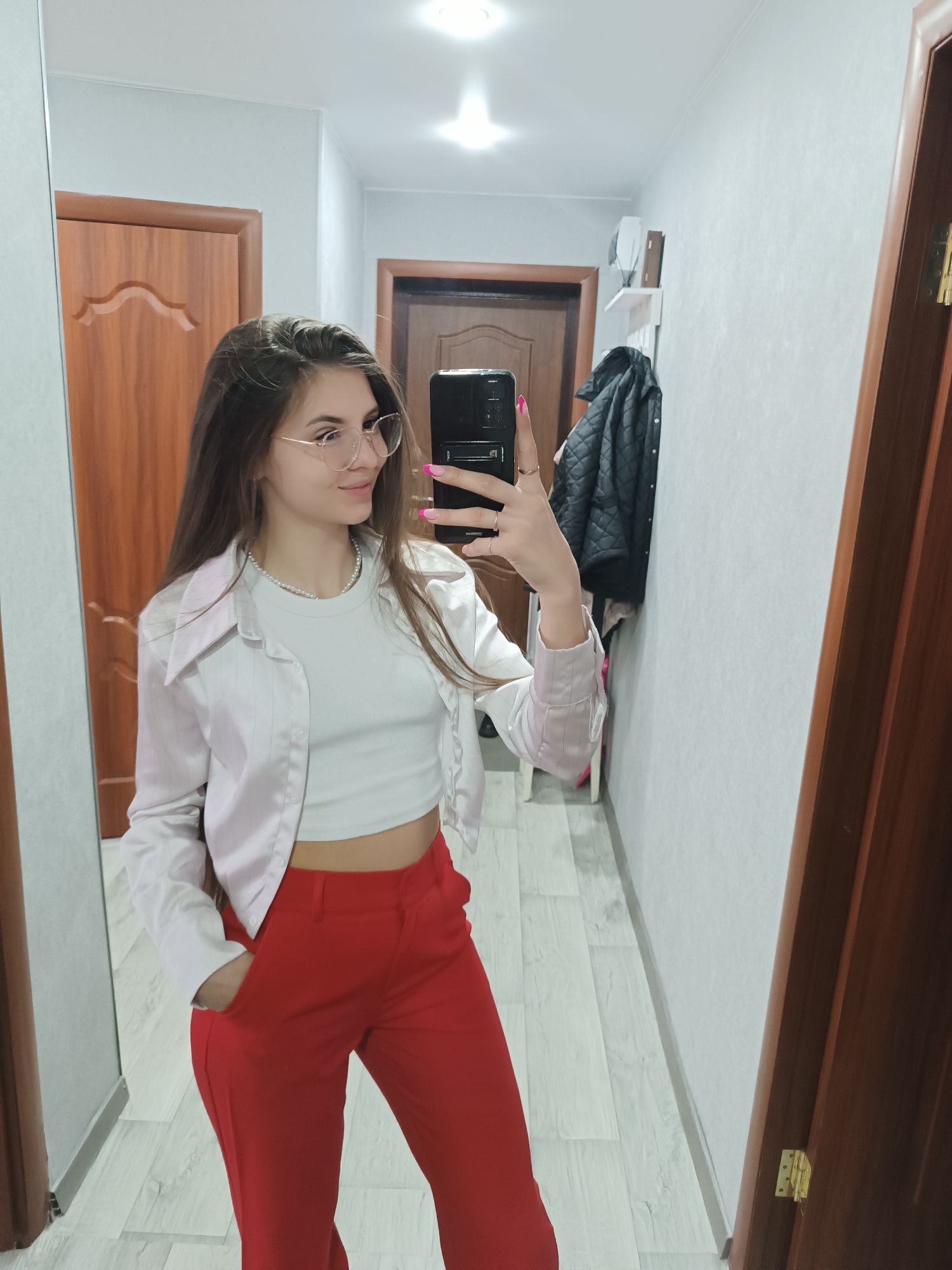 An indoor mirror selfie depicts a young adult female standing in a hallway within a residence in the city of Unknown, Unknown. The subject has long, straight, dark brown hair, wears round clear-framed eyeglasses, a white ribbed crop top, an open light-colored satin-like long-sleeved shirt, and red trousers. A single strand pearl necklace is visible, as are multiple rings on her left hand. She holds a black smartphone with her left hand, positioned to capture the image, while her right hand is in the pocket of her trousers. The reflected environment includes light-colored laminate flooring, light grey walls, and two brown wooden doors. A white floating shelf containing several items and a dark quilted jacket hanging below it are visible on the wall. Overhead lighting is present.