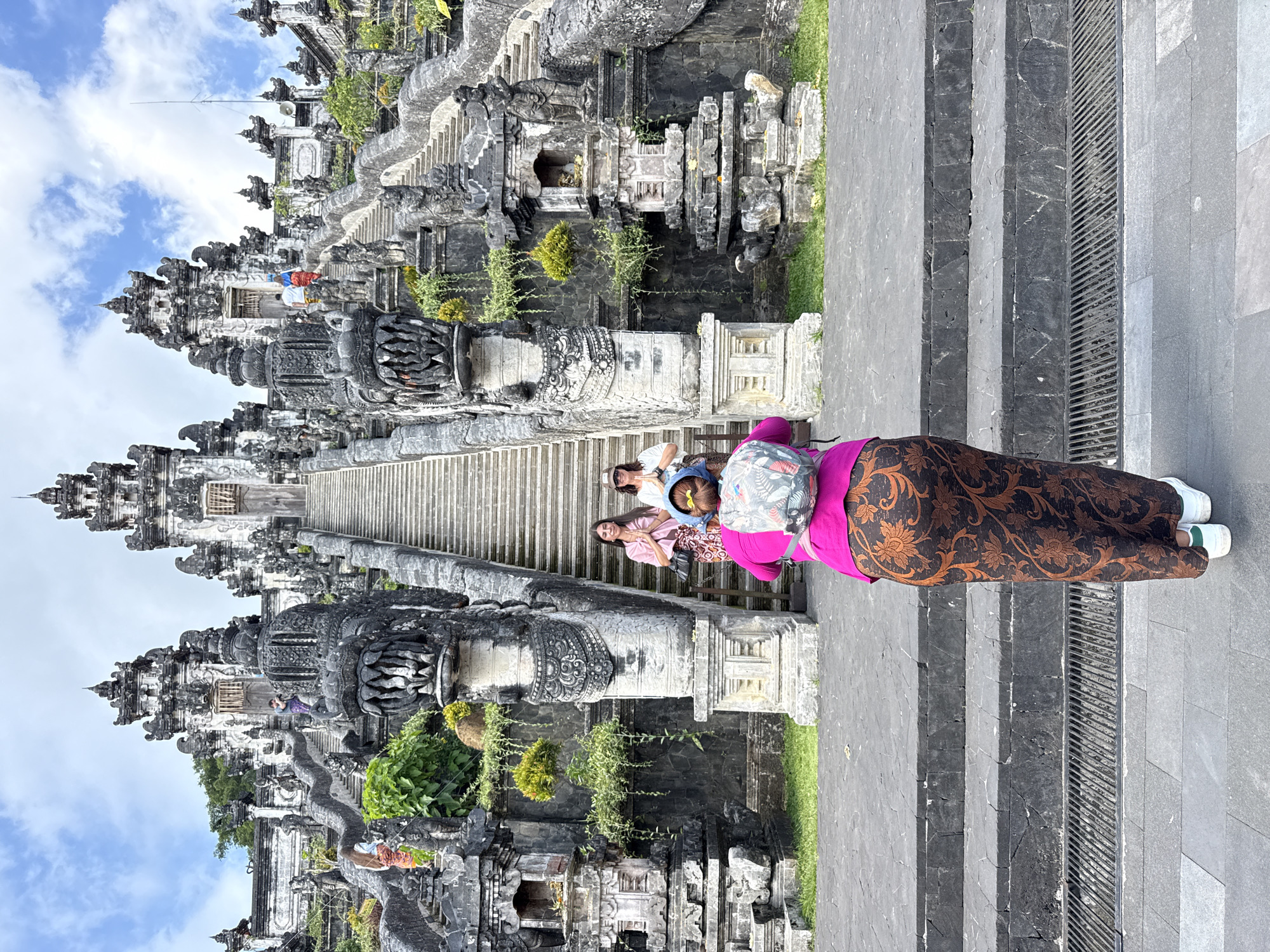 The image depicts the entrance to a Balinese temple, likely in Abang, Indonesia, characterized by its ornate stone architecture. In the foreground, a person is viewed from behind, wearing a bright pink top and a long, patterned brown skirt, with a backpack on their shoulders. They are facing a grand stone staircase that ascends to the temple structure. Two other individuals are positioned on the staircase, one in a light pink top and the other in a white top and a patterned skirt, both with their hands together in a prayer-like gesture. The staircase is flanked by intricate stone carvings, including dragon-like sculptures at its base. The temple itself features multiple levels and towers, constructed from dark, weathered stone, with lush greenery clinging to its sides. The sky above is partly cloudy, with patches of blue visible. The scene conveys a sense of cultural and spiritual significance associated with the temple complex.