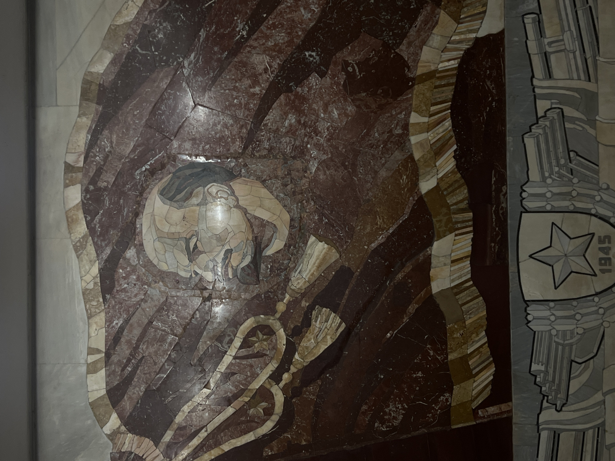 A close-up shot shows a marble mosaic design embedded in a wall. The mosaic features an image that appears to be partially figurative, possibly depicting a human figure or symbolic representation. To the right of the main mosaic, a separate design includes a five-pointed star and the number "1945". The marble mosaic is constructed of varied brown, white, and tan stones. The location is Unknown, Unknown.