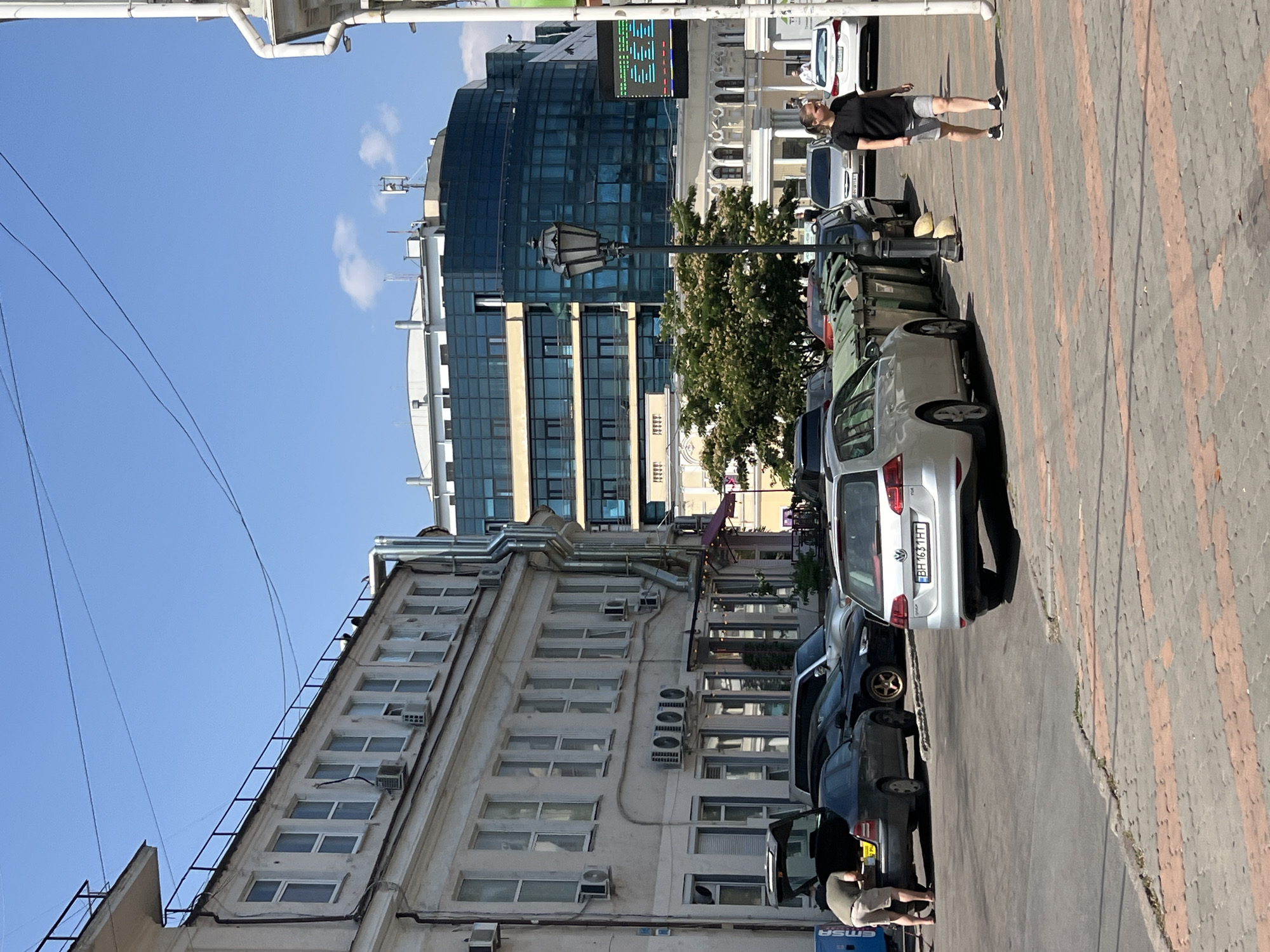 In Odesa, Ukraine, on a sunny day, a street scene unfolds in front of a multi-story building with large windows. Several cars are parked on the asphalt road, some with their trunks open. A person is seen interacting with the open trunk of a dark-colored sedan. Across the street, large green garbage bins are lined up. A woman in athletic shorts and a black t-shirt walks along the patterned sidewalk. In the background, a modern glass-facade building rises above older structures. The sky is clear and blue with a few wispy clouds.