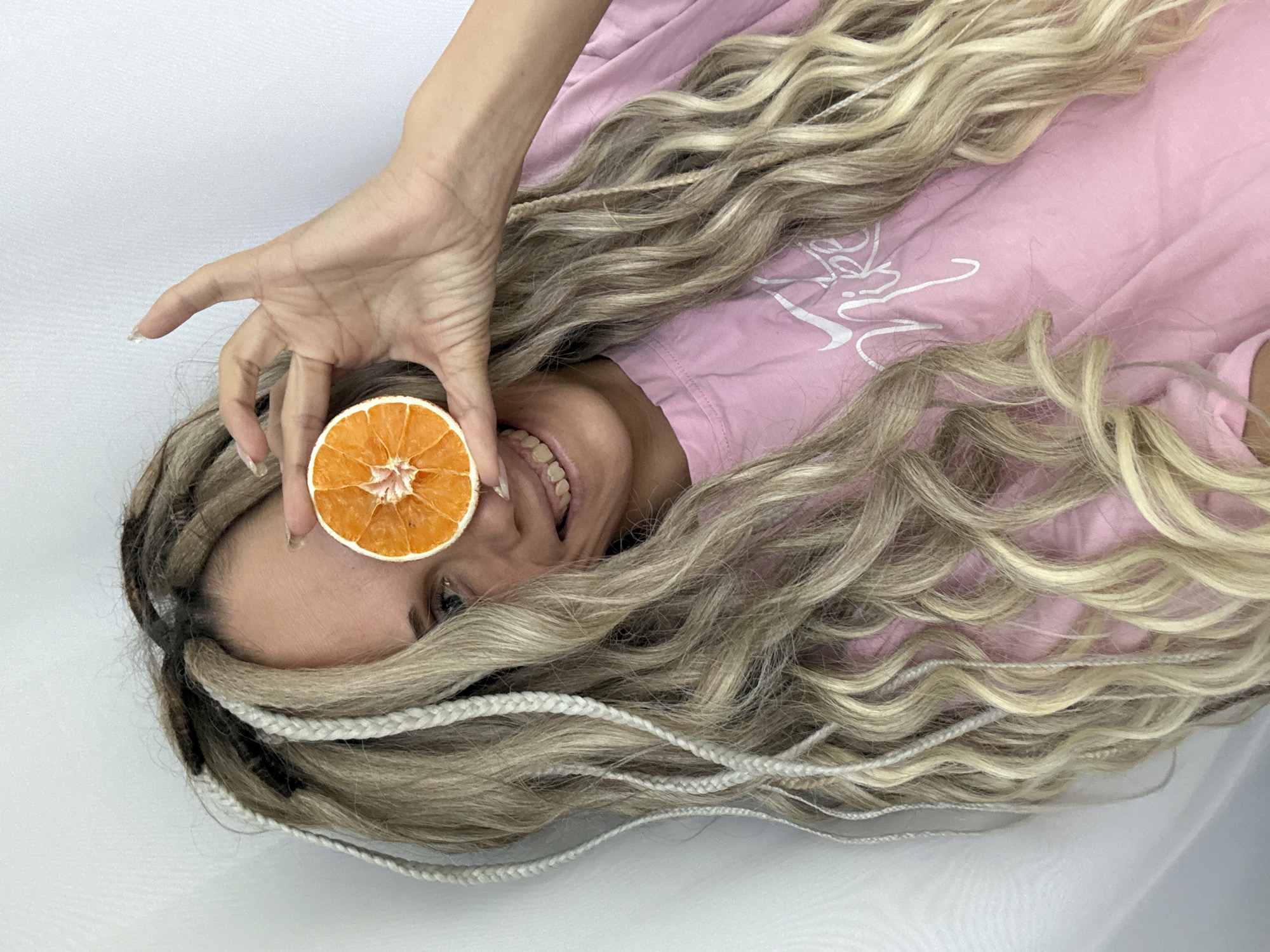 A fair-skinned woman with long blonde and brown hair is lying on her back, smiling and holding a dried orange slice in front of her left eye. She is wearing a pink shirt. The background is white. Rusanovo, Russia is not discernible in the image.