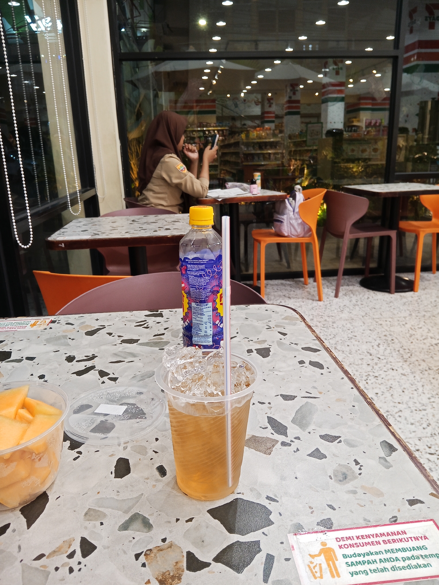 The image depicts a table in what appears to be a cafe or convenience store with outdoor seating. In the foreground, there is a plastic cup filled with ice and a light brown beverage, likely iced tea, with a straw. To the left of the cup, a clear plastic container holds sliced yellow fruit, possibly cantaloupe. A bottle of a purple beverage with a yellow cap is also visible on the table, with a straw extending from the cup. In the background, through a large window, shelves of products are visible, suggesting the establishment is a convenience store. A woman wearing a brown hijab and a uniform is seated at a table in the background, looking at her phone. Several brightly colored chairs are arranged around tables. A small sign on the table in the foreground features an orange graphic of a person throwing trash into a bin, with text in Indonesian that reads "DEMI KENYAMANAN KONSUMEN BERIKUTNYA Budayakan MEMBUANG SAMPAH ANDA pada tempat yang telah disediakan" which translates to "FOR THE CONVENIENCE OF THE FOLLOWING CONSUMERS, Cultivate THROWING YOUR TRASH in the designated place." The setting is consistent with a 7-Eleven store, which are present in Lubuklinggau, Indonesia.