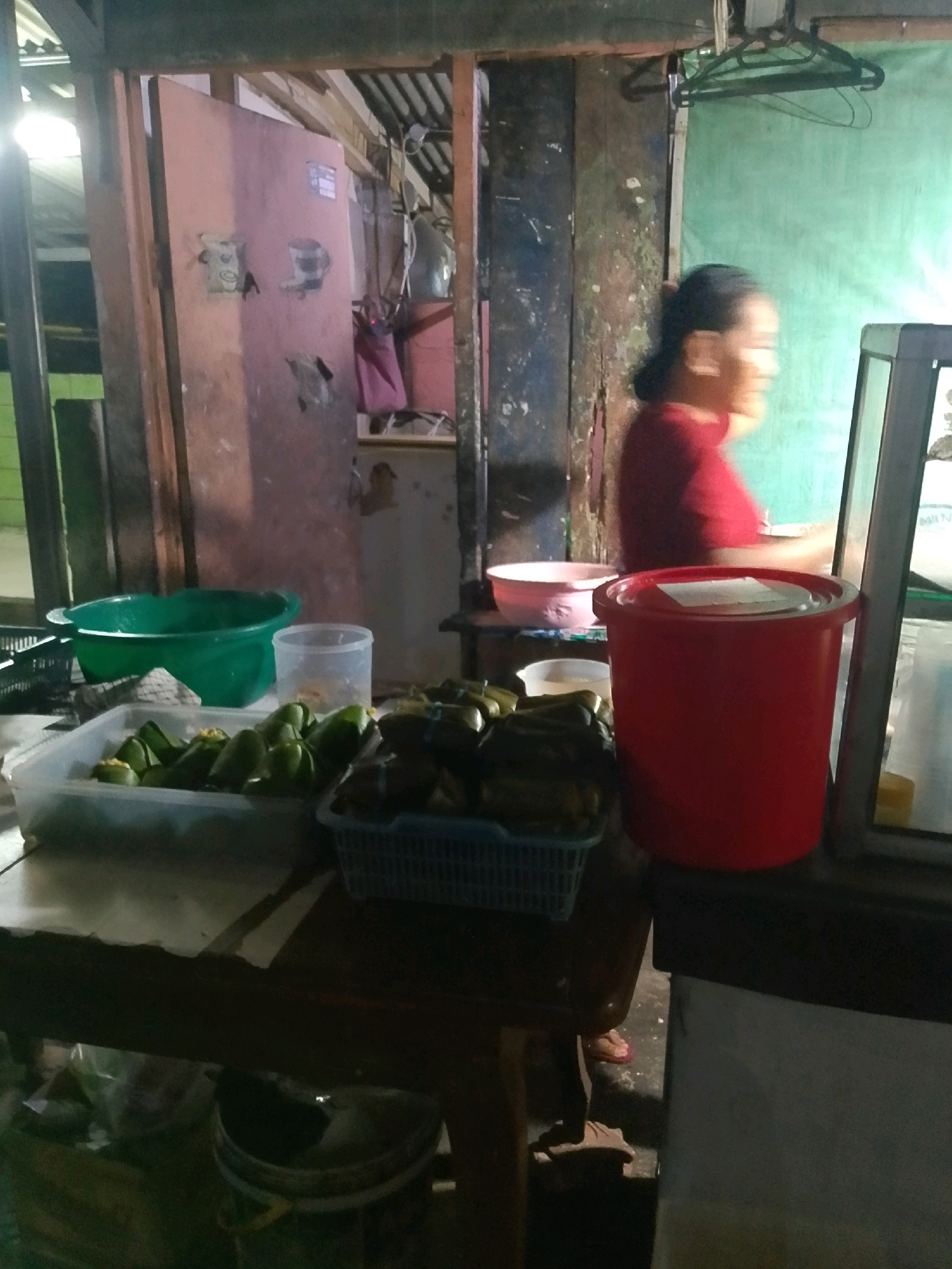 A woman in a red shirt stands in what appears to be an outdoor food stall in East Jakarta, Indonesia. She is positioned to the right of the frame, with her profile facing left, and is blurry due to motion. In front of her is a large red plastic container. To the left of the red container, a green plastic basin is visible, filled with water. Next to the green basin is a clear plastic container holding several green, banana-leaf-wrapped food items. Further to the right, a blue plastic basket contains more of the same food items. The stall is constructed from wood and corrugated metal, and the background is obscured by a green tarp and a pink door. The overall lighting is dim, suggesting it is either evening or the stall is covered.