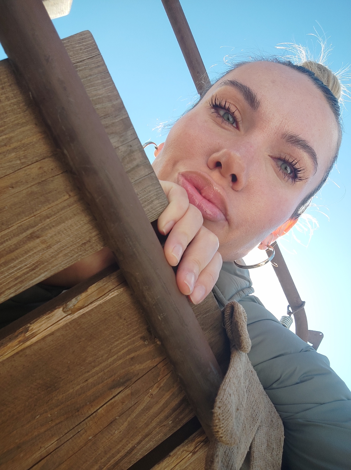 A woman poses close to the camera from a low angle, holding onto a weathered, brown wooden structure. She has her blonde hair in a bun and wears gold hoop earrings, visible against the bright blue sky in the background. She wears a light green puffer jacket, and a pair of light brown knitted gloves hang from her jacket. The location is not specified within the image. The city of Votkinsk, Russia, cannot be confirmed as the location.