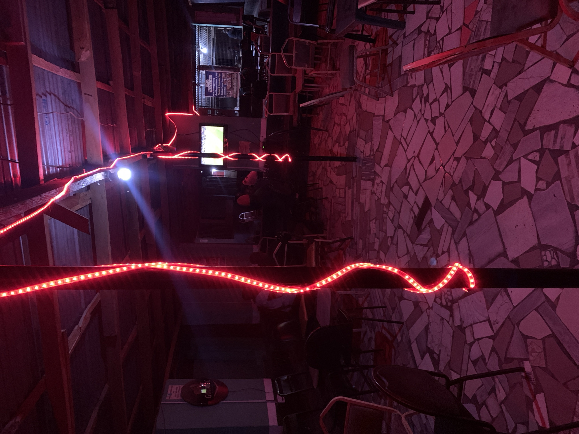 The image depicts the interior of a dimly lit bar or restaurant near New Stadium, Nigeria. The walls are constructed with a mix of wood and corrugated metal, and the floor is covered in irregular stone tiles. Red LED strip lights run along the upper sections of the wall and ceiling, providing the primary source of illumination. Tables and chairs are visible, mostly unoccupied. A television screen can be seen in the background. A sign listing kitchen items is visible on a nearby wall. A branded item labeled "Gulder" is visible on a table top near the bottom of the image. The scene suggests a casual, possibly late-night, setting.