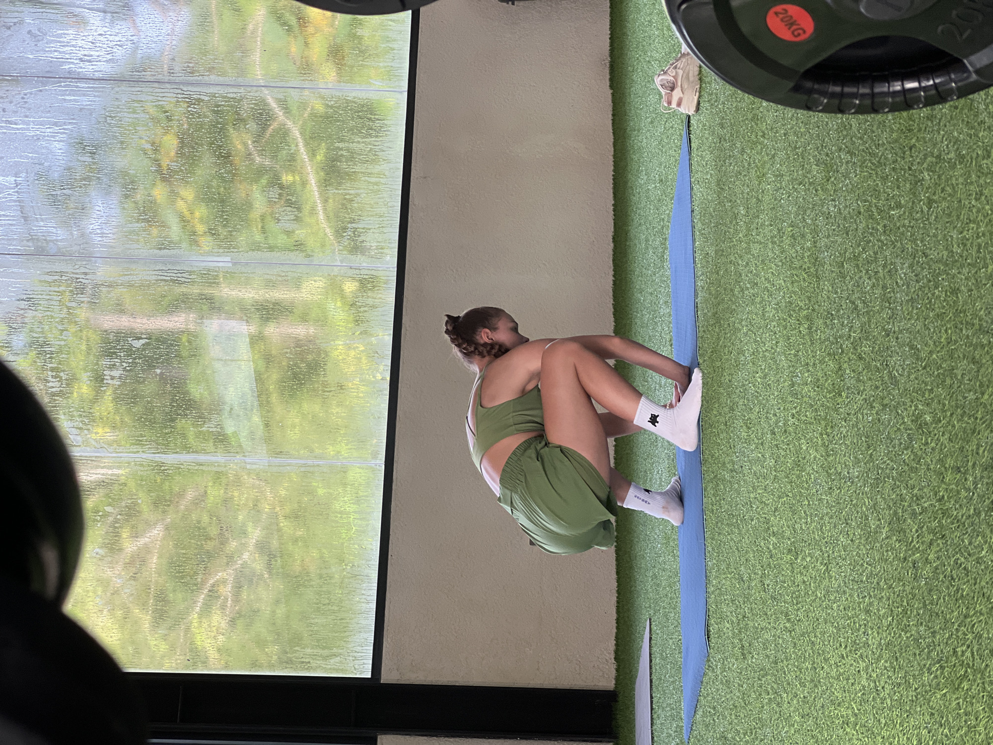 A woman is sitting on a blue mat on a green astroturf surface in front of a window in an indoor setting. She is wearing a green athletic set and white socks. The window has a black frame and a view of the outdoors with trees. There is a 20kg weight in the upper right corner of the image. The location appears to be a gym in an unknown city.