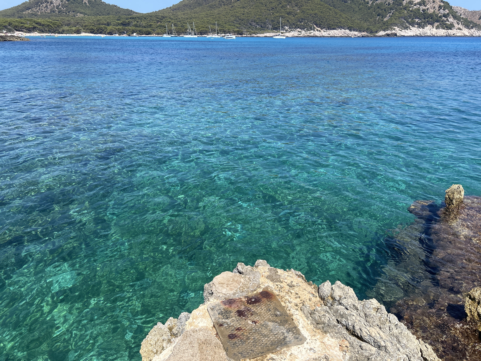 This media file is a landscape photograph taken at Cala Lliteres, Spain. The image captures a serene coastal scene with crystal-clear turquoise water in the foreground, transitioning to a deeper blue further out. The seabed is visible beneath the shallow water, with rocks and hints of greenery.

In the mid-ground, several sailboats are anchored in the bay, their white sails contrasting with the blue of the sea and sky. The background features a verdant, tree-covered hillside that slopes down to the coastline. A small beach with scattered people and beach umbrellas is visible along the shore, suggesting a popular recreational spot. The rocky shoreline in the foreground is textured and appears to be dry. A square metal plate with a grid pattern, showing signs of rust and wear, is embedded in the rocks at the bottom center of the image.

The lighting suggests it is a bright, sunny day, possibly late morning or early afternoon, with the sun casting reflections on the water's surface. The overall mood is peaceful and inviting, conveying a sense of a warm, leisurely vacation day at the beach. There is no visible text in the image.