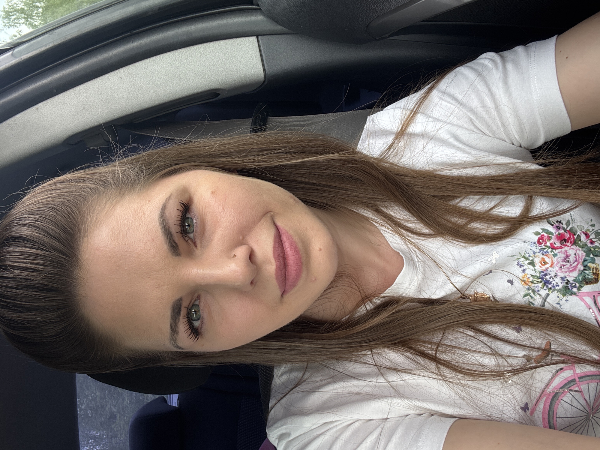 A woman with long brown hair, green eyes, and light skin is taking a selfie in a car. She is wearing a white t-shirt with a floral design. The car's interior is visible in the background, including the dashboard and headrest. The location is not identifiable as Shevlyakovo, Russia from the provided image.