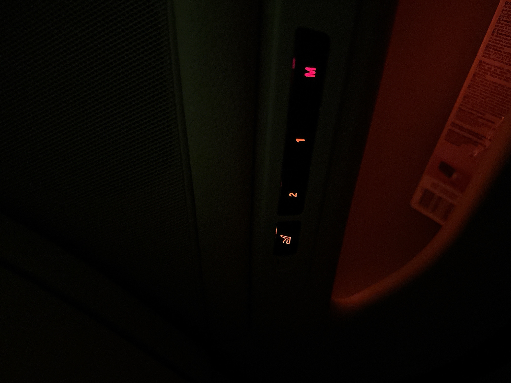 The image captures a dark interior scene, likely within a vehicle at night or in low-light conditions. The primary subjects are a vertical panel of illuminated controls or indicators and a textured surface.
On the dark central panel, a series of symbols glow. From top to bottom, these include a red letter 'M', a red numeral '1', an orange numeral '2', and an orange icon that appears to represent a seat with an arrow or a hand pointing. These symbols are brightly lit against the dark background.
To the left of these controls, a dark, finely gridded or woven texture is visible, likely the vehicle's headliner or another interior surface. On the far right, a lighter-colored object, possibly a sun visor or a piece of paper/card, is partially visible and illuminated by a warm, reddish-orange light. This object displays small, densely packed text, which is largely unreadable due to blur and lighting, but appears to be instructional or warning labels. A fragment that might be "гра" (part of a word in Cyrillic) is faintly discernible in the top right section of this object.
There is no discernible activity taking place; the image is a static shot. The overall lighting suggests a dark environment, emphasizing the functional glow of the vehicle's interior controls.