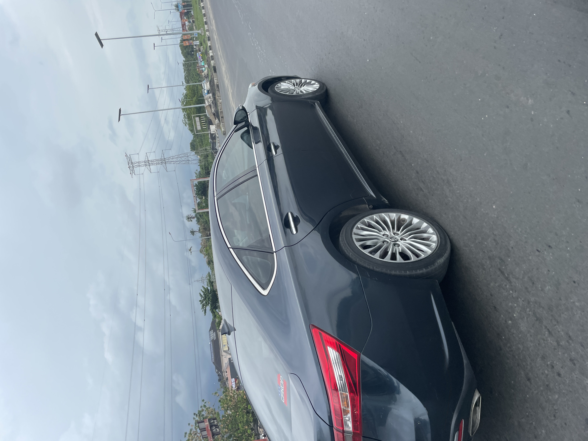 The image shows a dark-colored Toyota sedan driving on a paved road in Igboefon, Nigeria. Overhead power lines and poles are visible on the left side of the frame, along with buildings and trees. The sky is overcast.