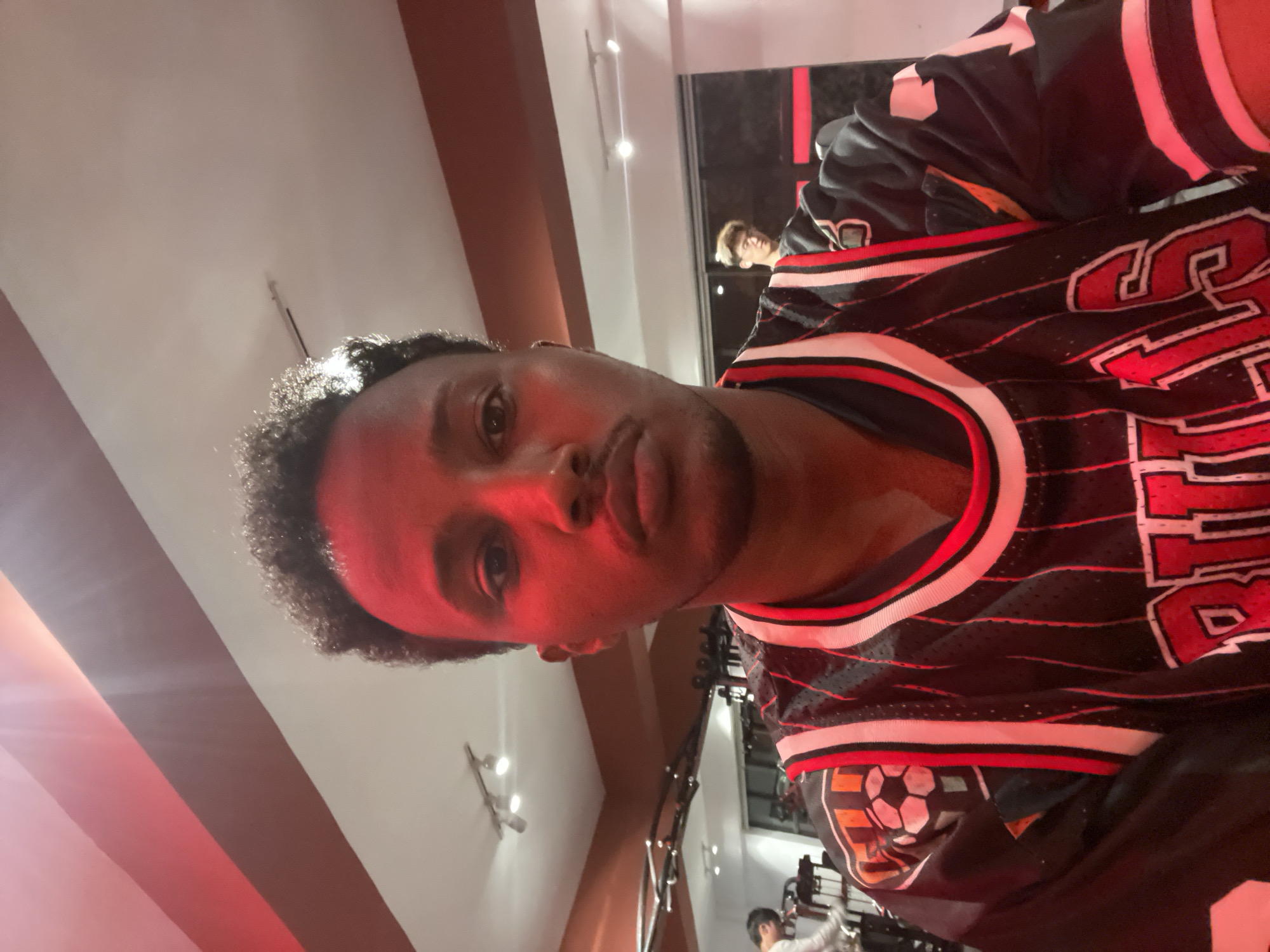 A man is wearing a black jersey with the word "BULLS" written in red and white letters, with black trim. The location is determined to be Chongqing High-tech Zone, China. Another person is visible in the background, seemingly in a mirrored reflection. The scene is indoors, potentially a gym or recreational facility, given the presence of exercise equipment.