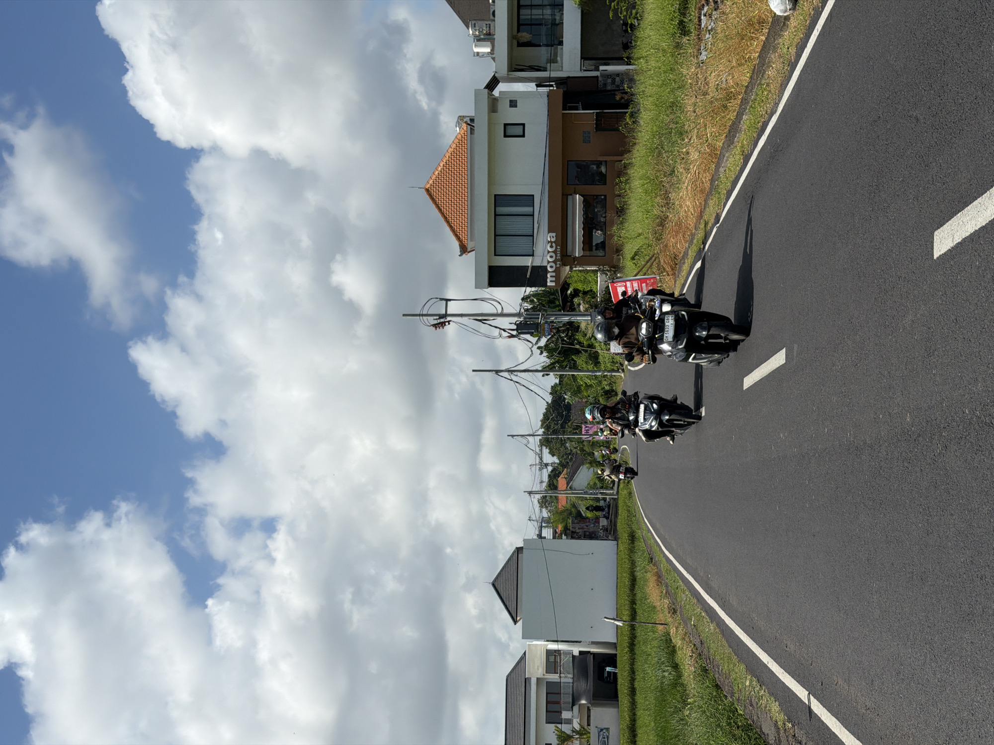 The image depicts a road in North Kuta, Indonesia, with two motorcycles driving along it. The road is asphalt and has a white line marking its edge. On either side of the road, there is a grassy area and buildings. The sky is partially cloudy. The motorcycles are occupied by people wearing helmets. "mooca" is visible on the storefront of a building.