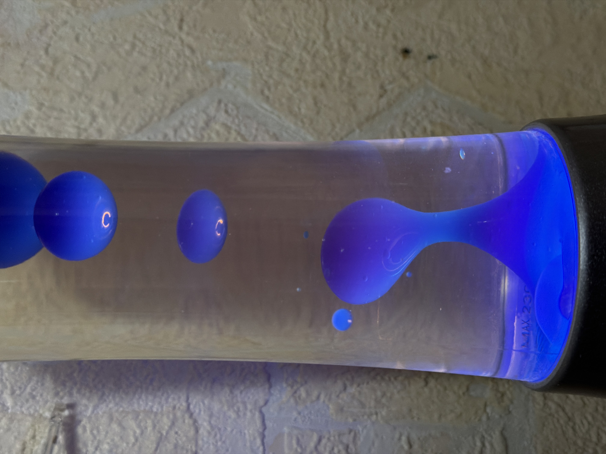 A horizontal, close-up shot captures an illuminated blue lava lamp positioned against a textured, light-colored wall. The cylindrical lamp, filled with a clear liquid, contains several bright blue wax blobs in various stages of movement and formation. On the left, two distinct spherical blobs of blue wax are visible, with reflections resembling the letter 'C' on their surfaces. Another smaller, spherical blob is positioned further right.
On the right side of the lamp, a large, amorphous mass of blue wax extends upwards, appearing to merge with the black cap or base of the lamp, which emits a vibrant blue light. A small, separate droplet of blue wax floats beneath this larger mass. The texture of the background wall suggests a rough surface, possibly brick or stucco, bathed in a soft, diffused light, contrasting with the intense blue glow of the lamp. Faintly visible through the glass and wax on the right side of the cylinder is text that reads "MAX 230". The image depicts the dynamic and mesmerizing activity of the lava lamp, characteristic of its operation. No people are visible, and the time of day is not discernible, though the artificial lighting suggests an indoor setting. The location is Izhevsk, Russia.