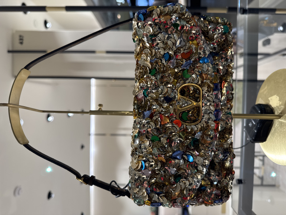 A Valentino brand shoulder bag is centrally displayed within a luxury retail boutique in Paris, France. The bag is rectangular, heavily embellished with iridescent silver sequins and numerous multi-colored faceted gemstones (including red, blue, green, and orange), featuring a prominent gold-toned "V" logo on its front flap. A slender, dark-colored strap with gold hardware is attached. The bag hangs from a brass-colored metal display stand with a circular base, upon which a small black security tag rests. The background reveals a bright, modern store environment with recessed ceiling lights, white walls or columns, and blurred display fixtures. A numerical label "437" is visible on a column in the upper right background. No individuals are present in the scene.