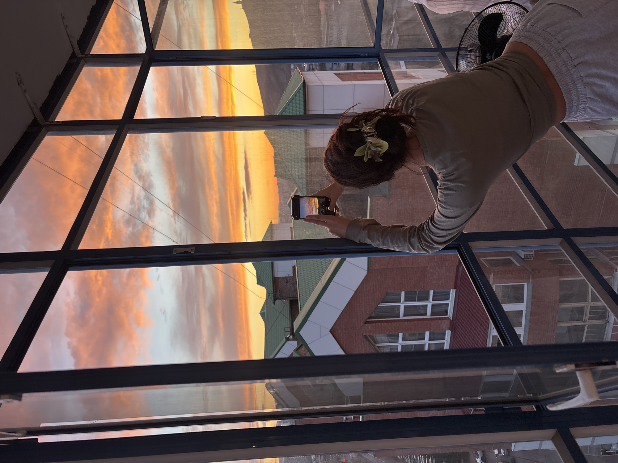 The image captures a vibrant sunset scene viewed from an indoor setting in Krasnoyarsk, Russia. The primary subject is a woman, seen from behind, with dark hair tied up and adorned with green accents, wearing a light-colored long-sleeved top and grey sweatpants. She is holding a black smartphone horizontally, actively taking a picture of the sunset through a large glass window.
The sky outside is ablaze with the dramatic colors of sunset, featuring prominent orange, yellow, and pink clouds that are vividly reflected in the window panes. The screen of her smartphone also displays the intense hues of the sunset she is capturing. Beyond the window and below the horizon, an urban landscape is visible, including buildings with red brick walls, green sloped roofs, and modern structures with white angular designs. A black electric fan is partially visible in the upper right portion of the frame, near the window. No discernible text is visible on any object. The scene conveys a moment of appreciation for the beautiful evening sky from an elevated, glass-enclosed space.