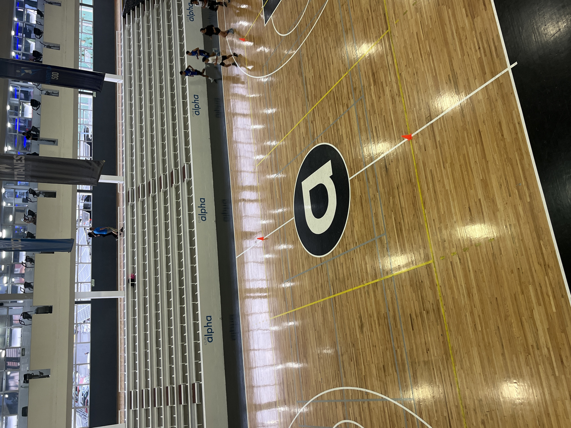 This image captures an indoor sports facility, specifically a basketball court with a polished wooden floor. The court is marked with standard white lines for basketball, along with additional yellow and grey lines, possibly indicating areas for other sports or training drills. A prominent black and white logo, resembling a stylized "D" or a graphic "alpha" symbol, is centered on the court.
Several individuals are visible on the court, dressed in athletic attire, with some wearing blue shirts and others dark clothing. They appear to be participating in a training session or warm-up, with some standing in lines, suggesting organized activity. Small orange cones are strategically placed on the court, commonly used as markers for drills.
To the left of the court, a long stretch of empty bleachers or tiered seating extends into the distance. These bleachers prominently display the text "club alpha" multiple times. Beyond the bleachers, large windows offer a view into another section of the facility, where various exercise machines like treadmills and stationary bikes are visible, indicating a broader fitness center.
Two large banners hang vertically from the ceiling in the background. One banner reads "JUVENTU 500," and the other displays "TITANES alpha." The facility is brightly lit, both by natural light filtering through the windows and by artificial interior lighting, suggesting it is daytime. The overall scene conveys an active and well-equipped athletic environment.
