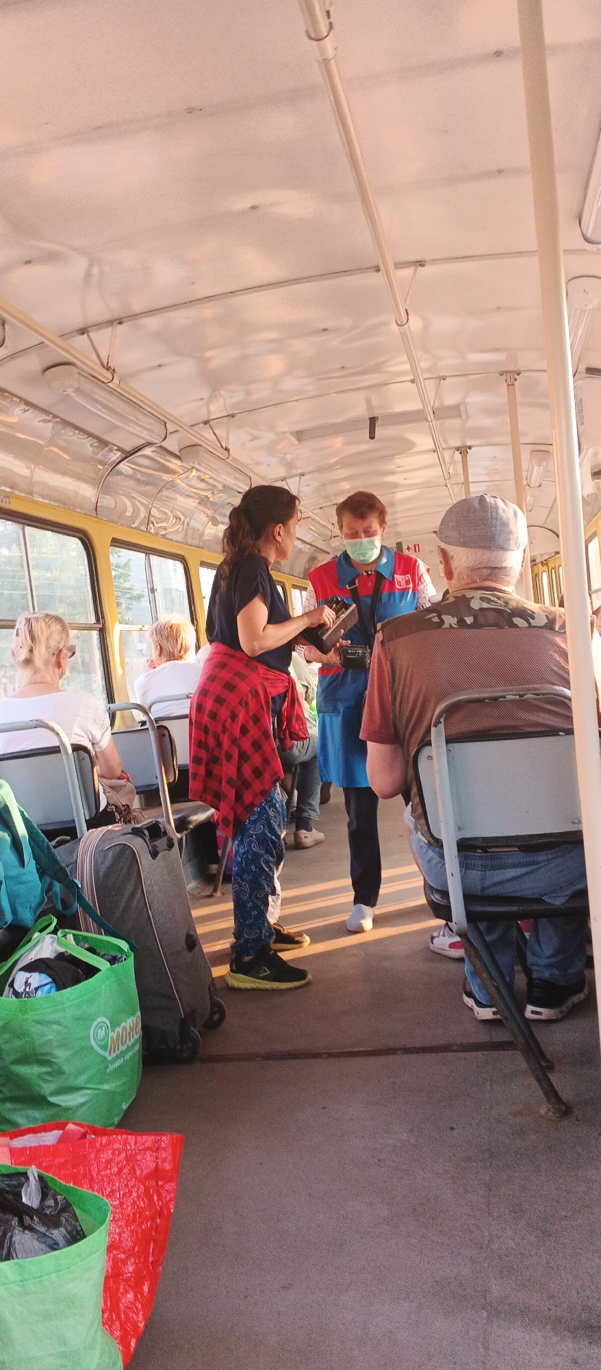 The image depicts the interior of a tram in Izhevsk, Russia, with several passengers visible. In the foreground, on the right, a man wearing a camouflage jacket and a grey cap is seated, facing away from the camera. To his left, a woman with dark, curly hair tied back is interacting with a man who is wearing a blue and red uniform, a light green face mask, and a lanyard with a device attached. The woman is wearing a dark t-shirt and a red and black checkered shirt tied around her waist, along with patterned trousers and black sneakers. She is holding a dark rectangular object, possibly a ticket validation device or a small electronic gadget. In the background, other passengers are seated in the tram, including two women on the left side, one with blonde hair and the other with grey hair. Luggage, including a grey wheeled suitcase and green and red shopping bags, is placed on the floor near the seated passengers. The tram's interior features yellow-tinted windows and overhead lighting fixtures, with a pale, possibly white or light pink, ceiling. The lighting suggests it is daytime, with sunlight filtering in.